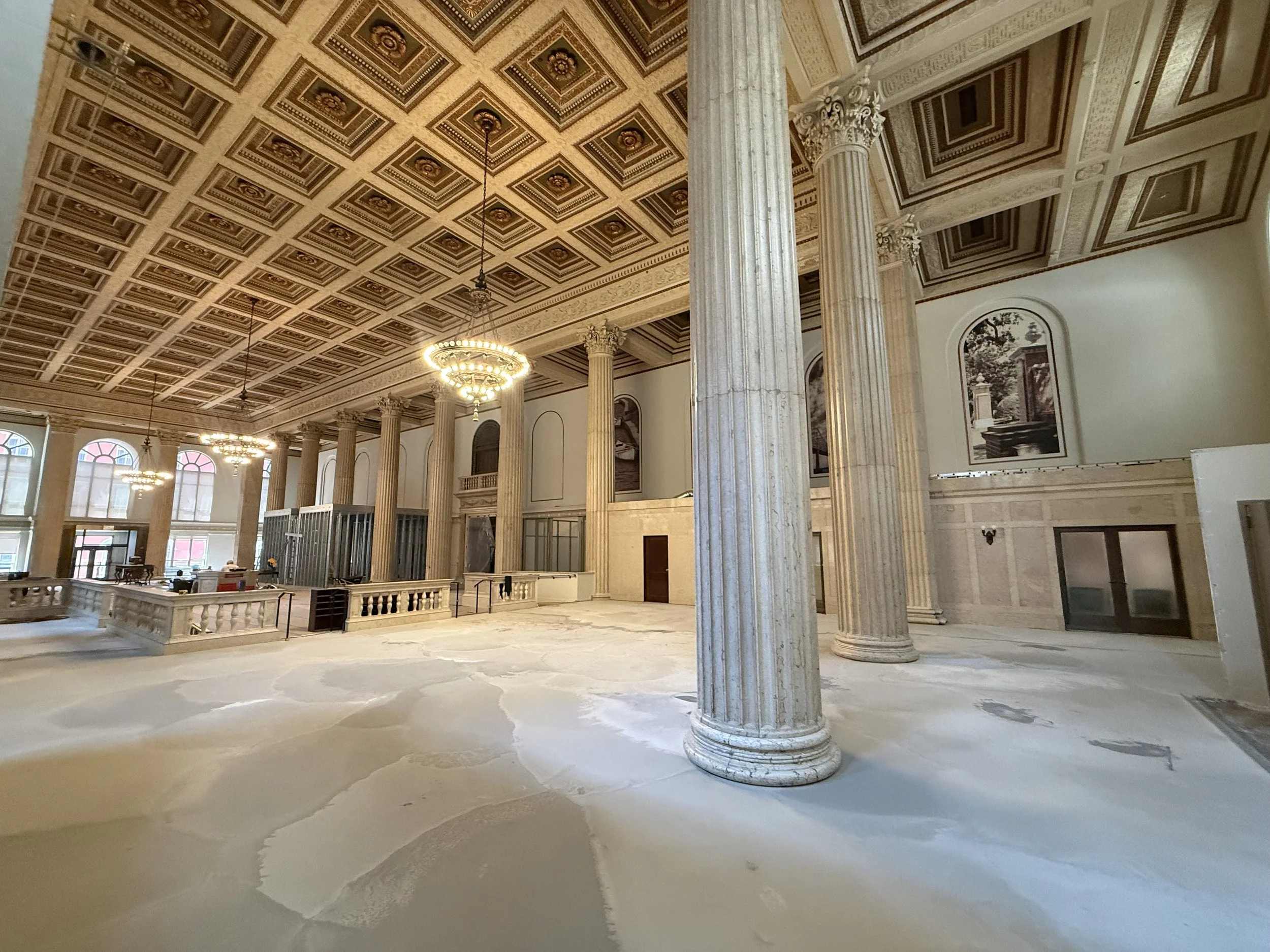 Interior of a grand hall with tall Greek-style columns, ornate coffered ceiling, and elegant chandeliers, that appears to be under construction or renovation.