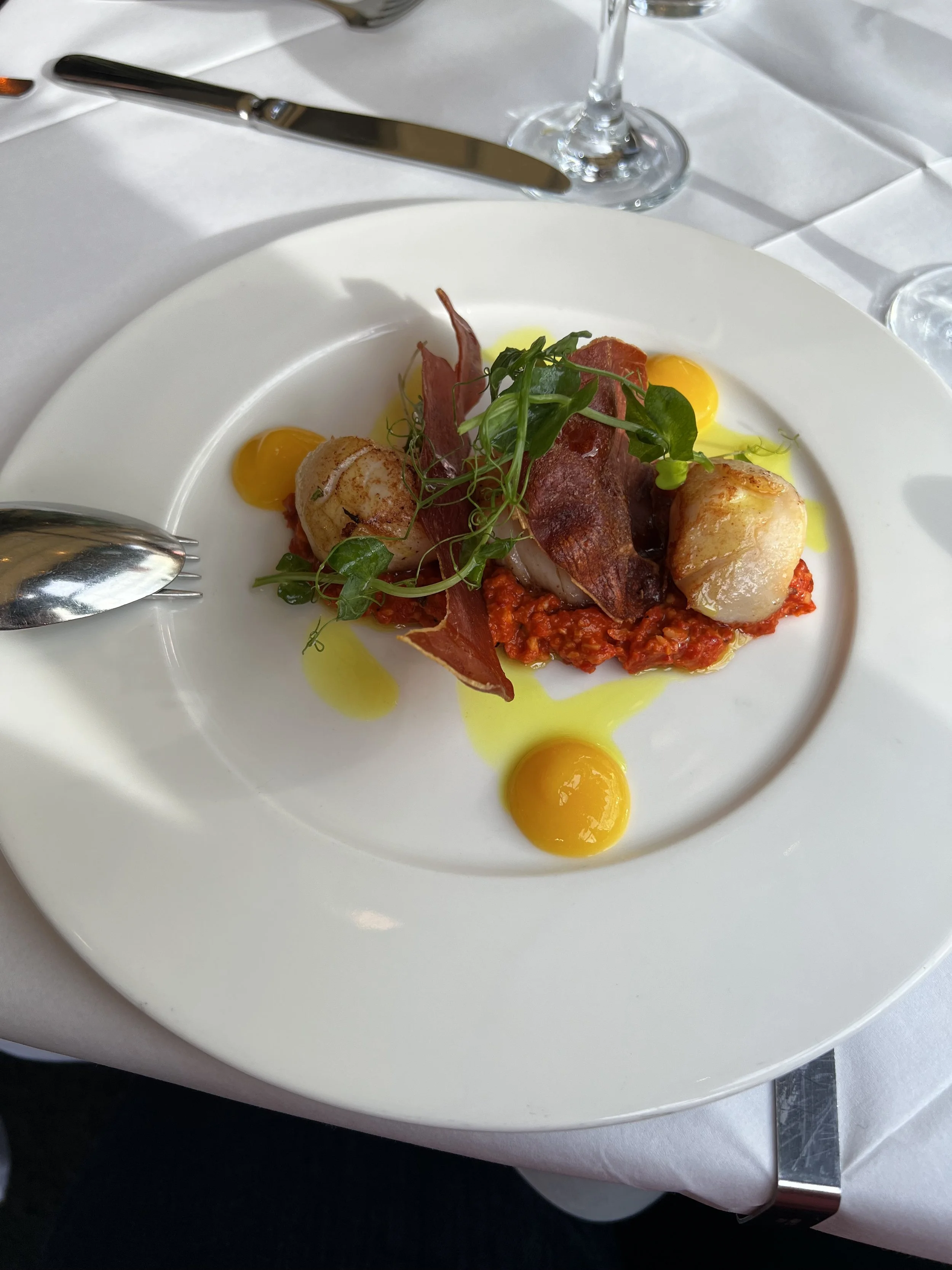 A plated gourmet dish featuring seared scallops, crispy bacon, microgreens, and a red pepper-based sauce, garnished with yellow oil drops and accompanied by a fork on a white plate.