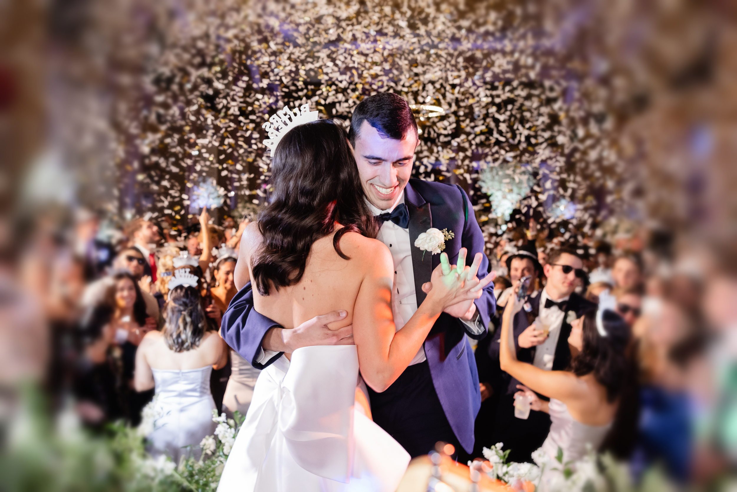 A newlywed couple dancing at their wedding reception with a background of confetti and guests. The bride has dark hair and is wearing a white gown, and the groom is wearing a tuxedo with a bow tie. They are smiling and holding hands.