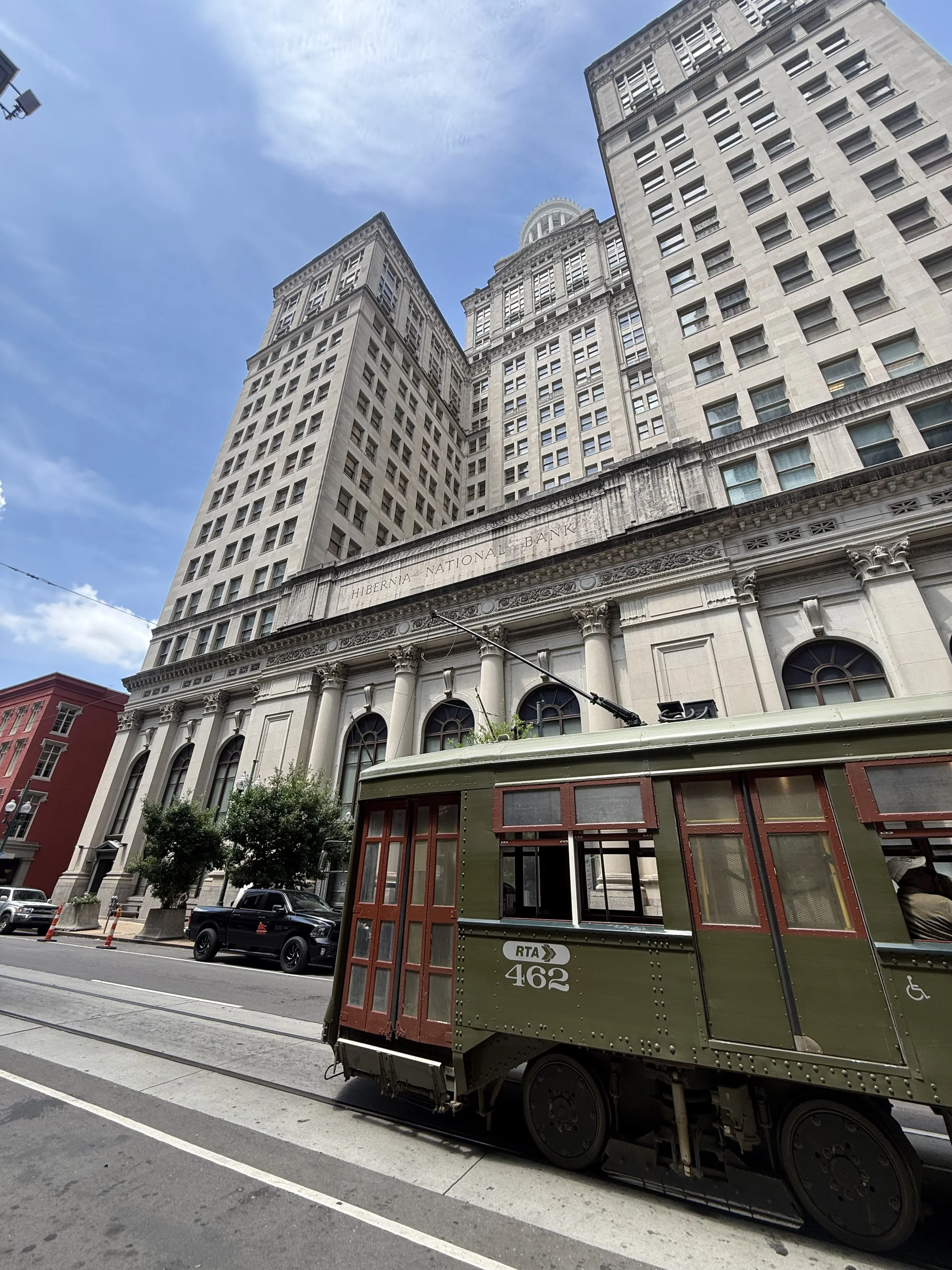 A vintage trolley painted green with red window frames, numbered 462, on city street tracks in front of large multi-story historic building, with modern cars parked nearby and a blue sky overhead.
