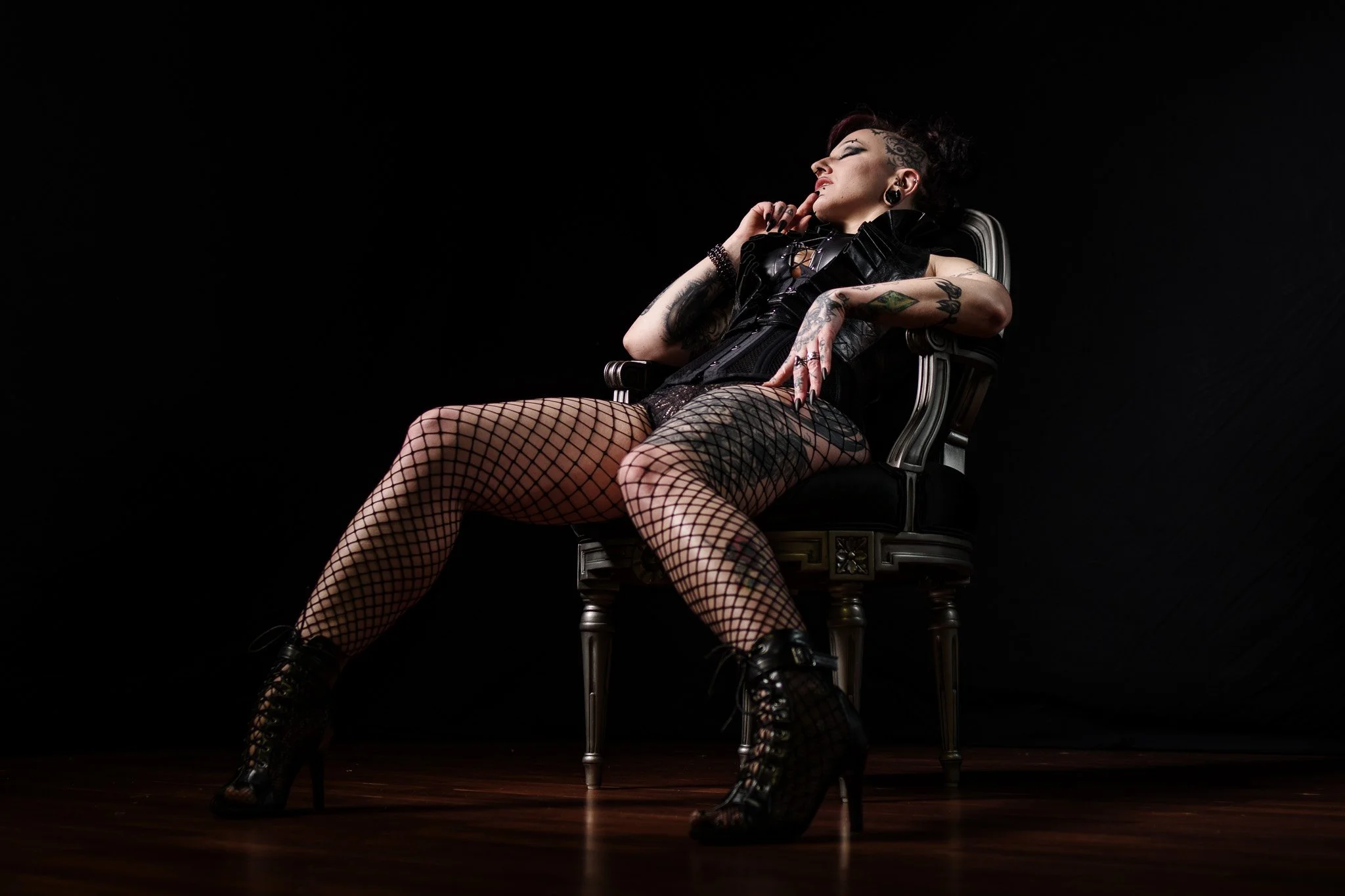 A woman with tattoos wearing fishnet stockings and platform boots sitting on a chair against a black background.