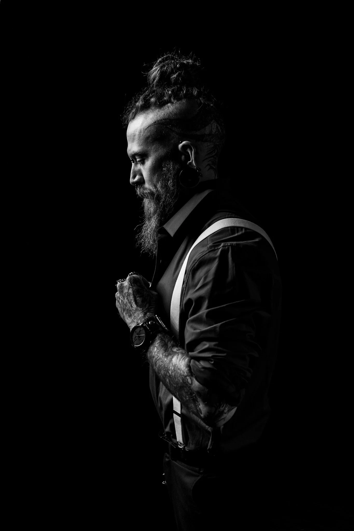 Black and white profile of a tattooed man with a beard and dreadlocks tied up in a high bun, wearing a jacket with suspenders, looking down with a serious expression, against dark background.