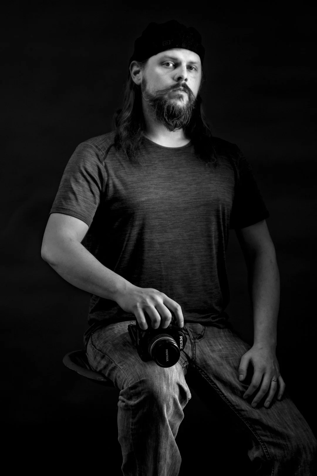 A black and white portrait of a man with long hair, a beard, wearing a black beanie and athletic shirt, holding a camera, sitting on a stool against a dark background.