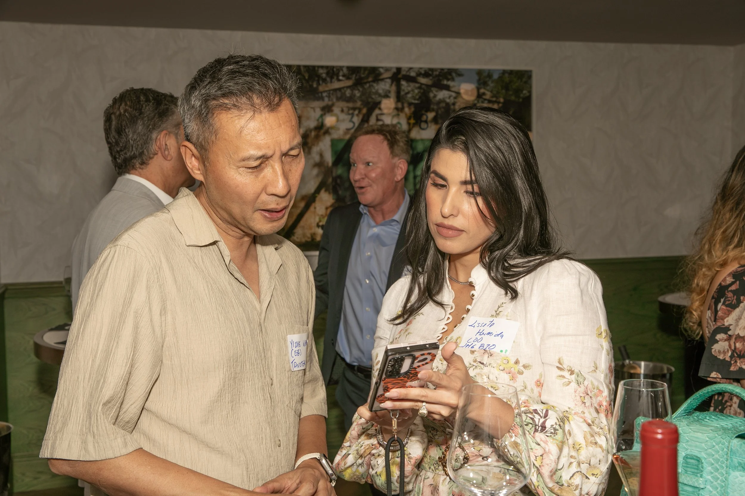 A group of people are gathered at a social event, with one woman showing her phone to a man. The woman has long dark hair and wears a floral blouse, while the man has short gray hair and wears a beige shirt. Several other people are in the background, engaged in conversation. There are name tags on the attendees indicating a networking or professional event.