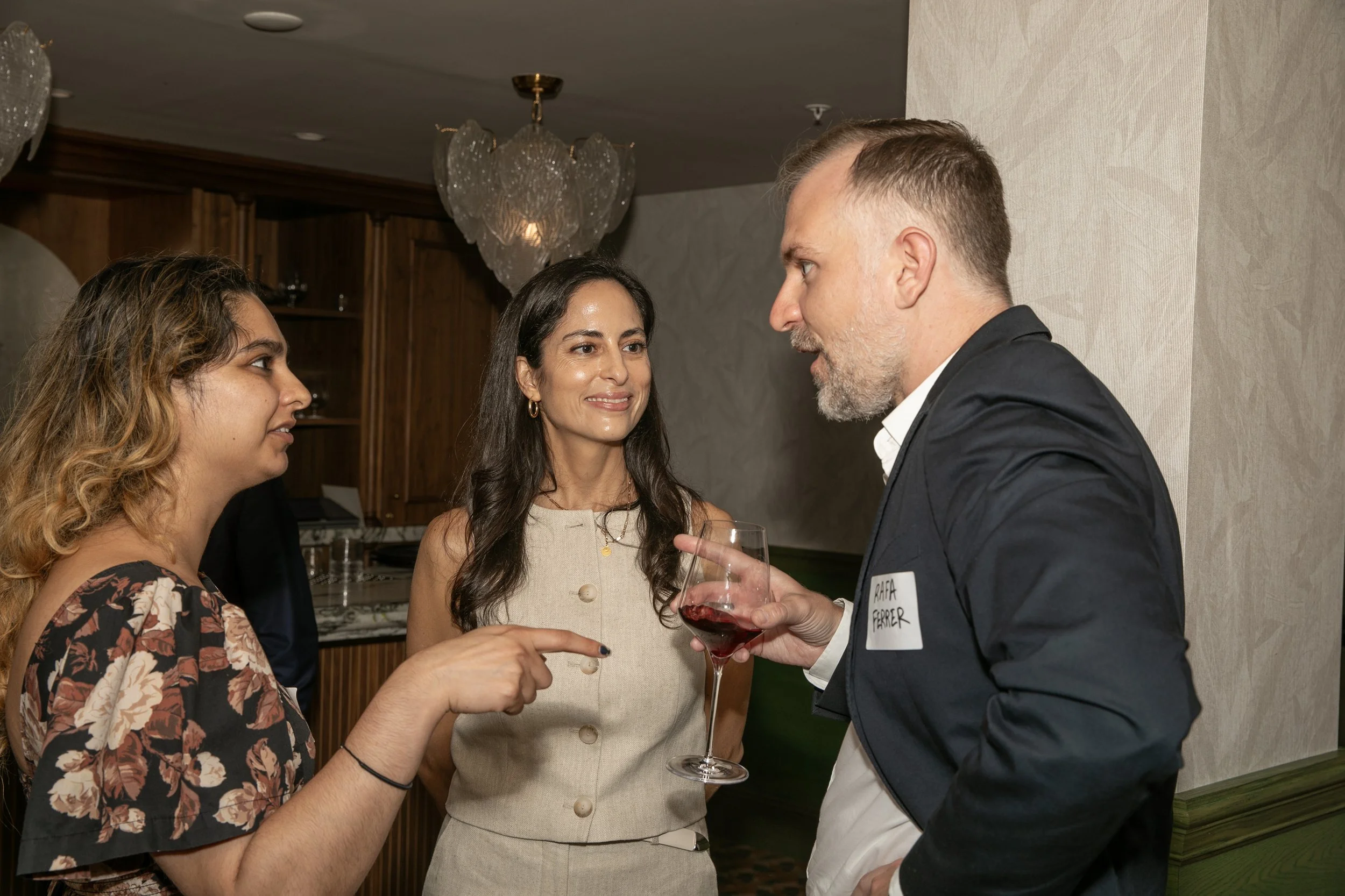 Three women and one man engaged in conversation at a social gathering, with the man holding a glass of wine. The woman in the middle is smiling and wearing a beige dress, and the woman on the left is wearing a floral top. The man is dressed in a suit jacket.