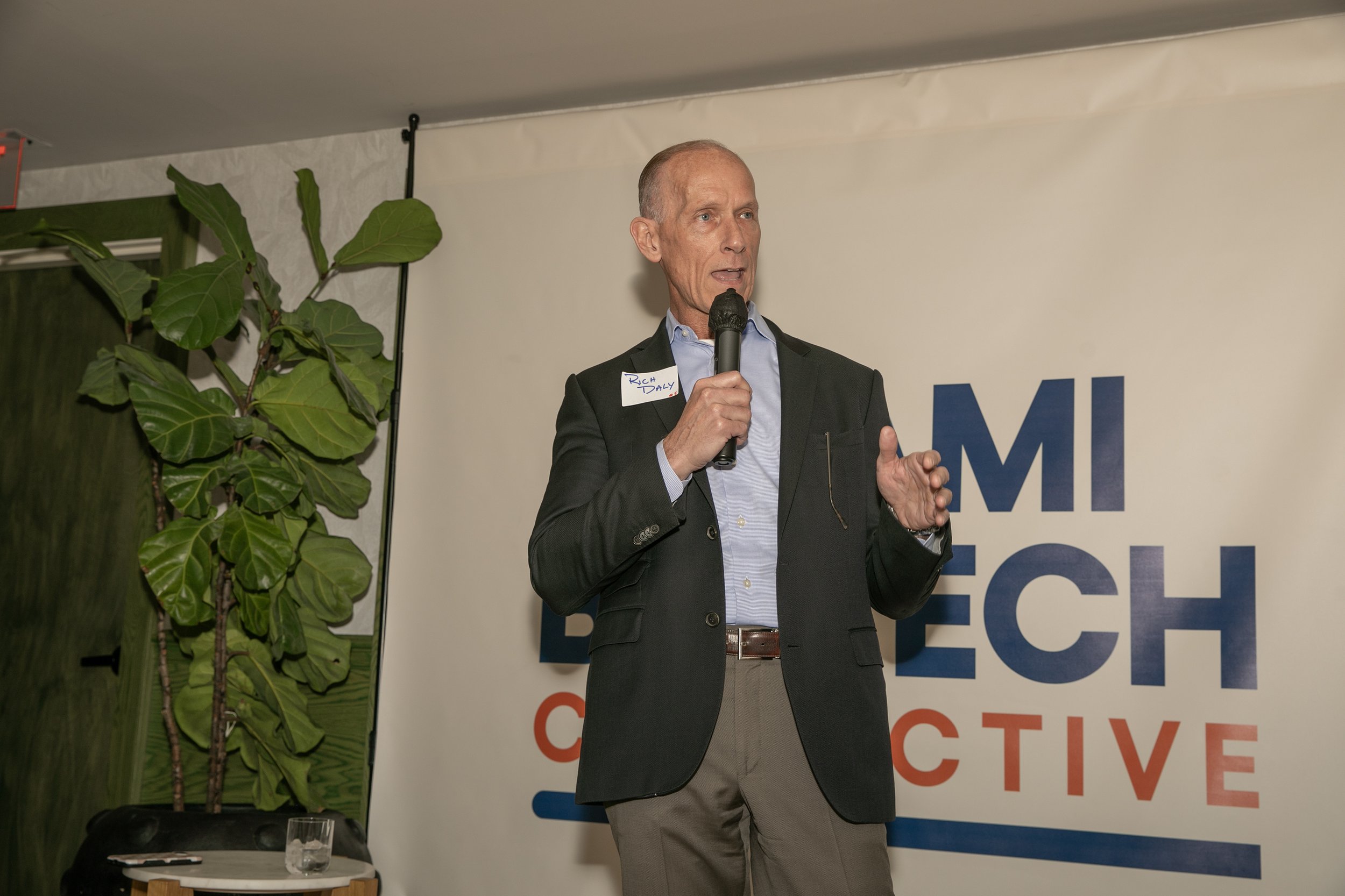 A man in a dark suit jacket and light shirt giving a speech while holding a microphone. There is a large plant to his left and a banner with partially visible text behind him.