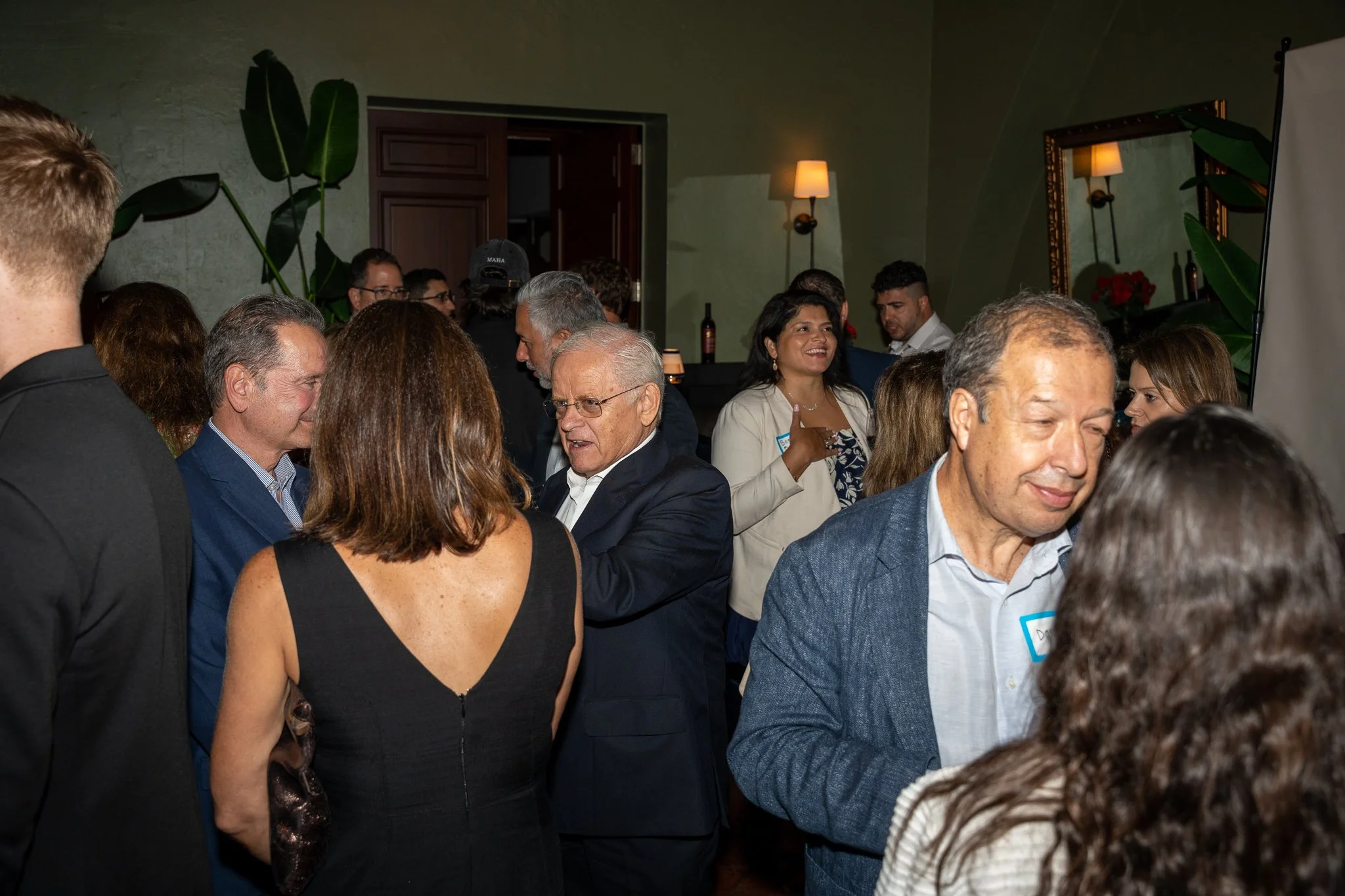 A crowd of people at a social event, engaged in conversation, indoors with green walls, plants, and warm lighting.