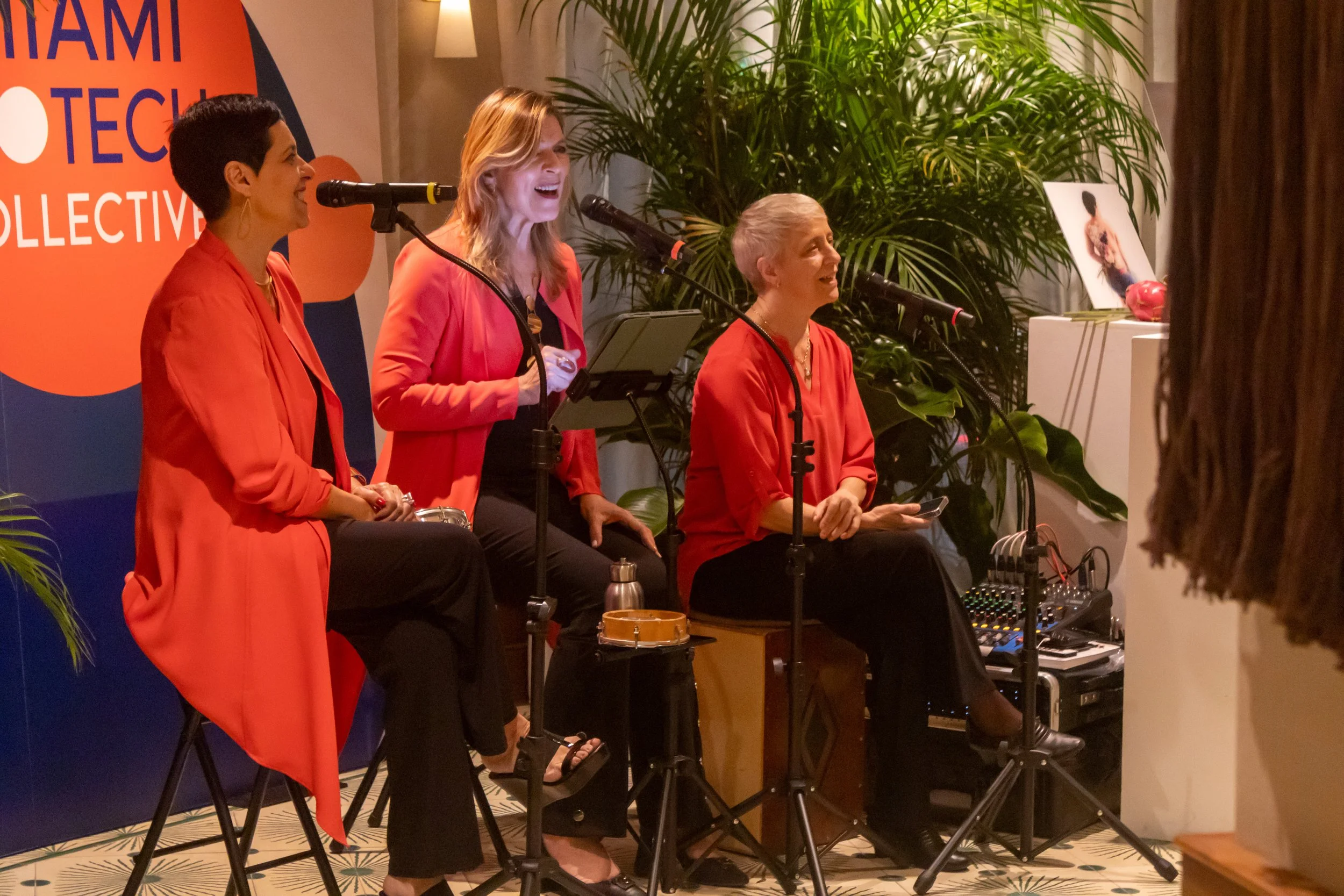 Four women are on stage, singing into microphones during a musical performance or panel discussion. They are dressed in red or coral outfits. The background features a large blue and orange banner and lush green plants.