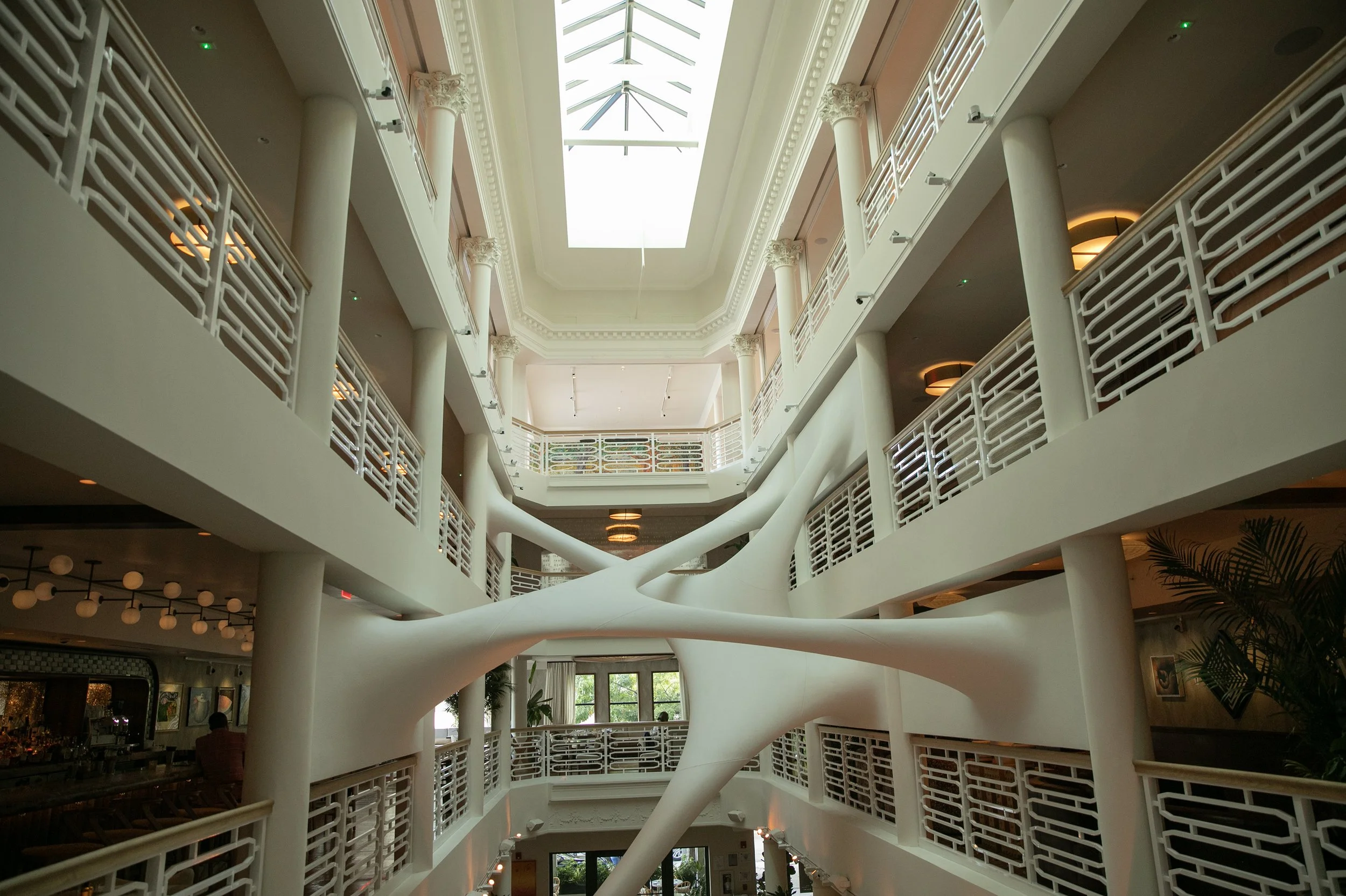 Interior of a modern multi-story building with a central open atrium, featuring white railings, columns, and a skylight ceiling.