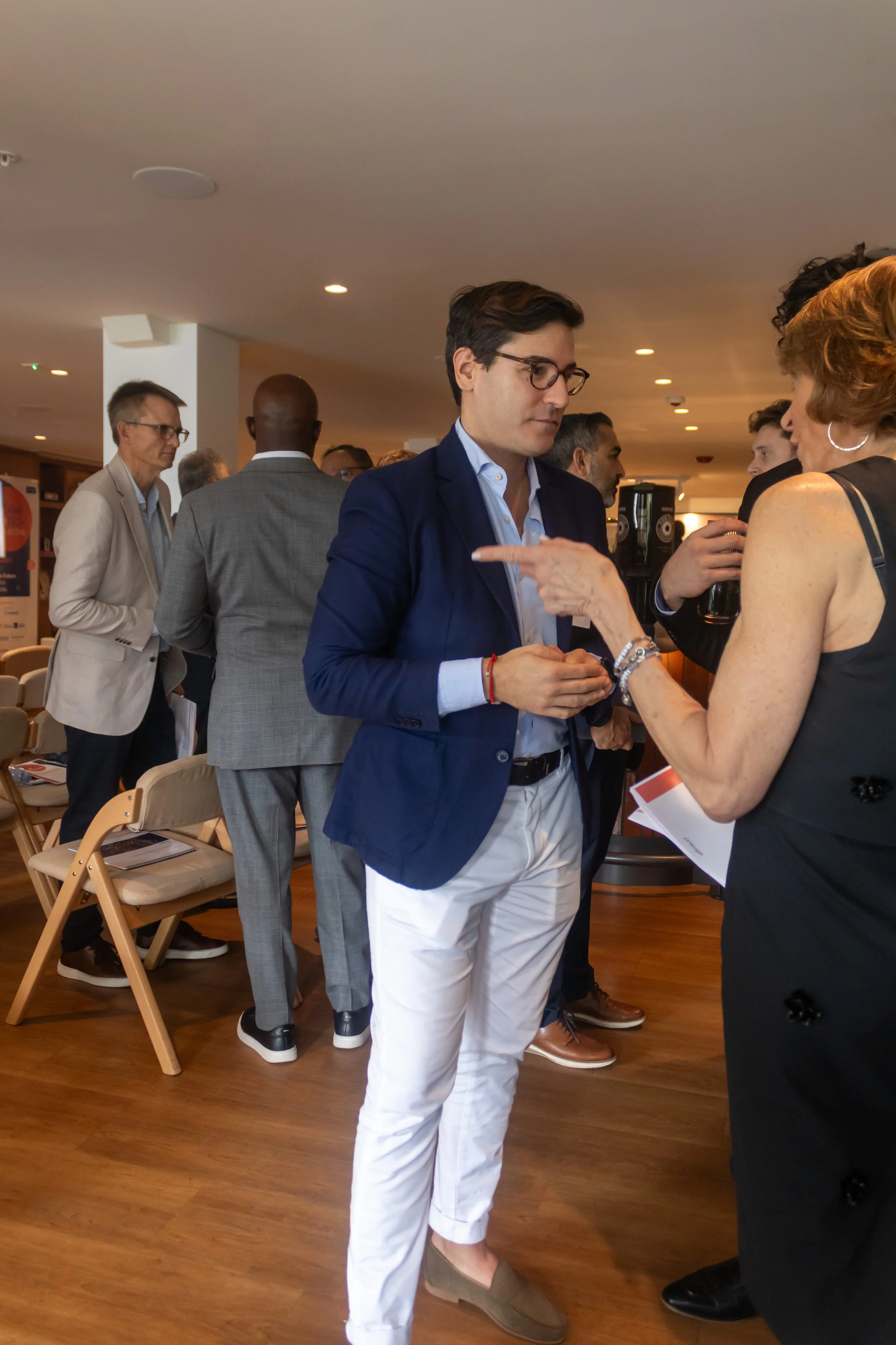 A group of people at a professional networking or conference event, talking and engaging with each other, in a room with wooden flooring and chairs.