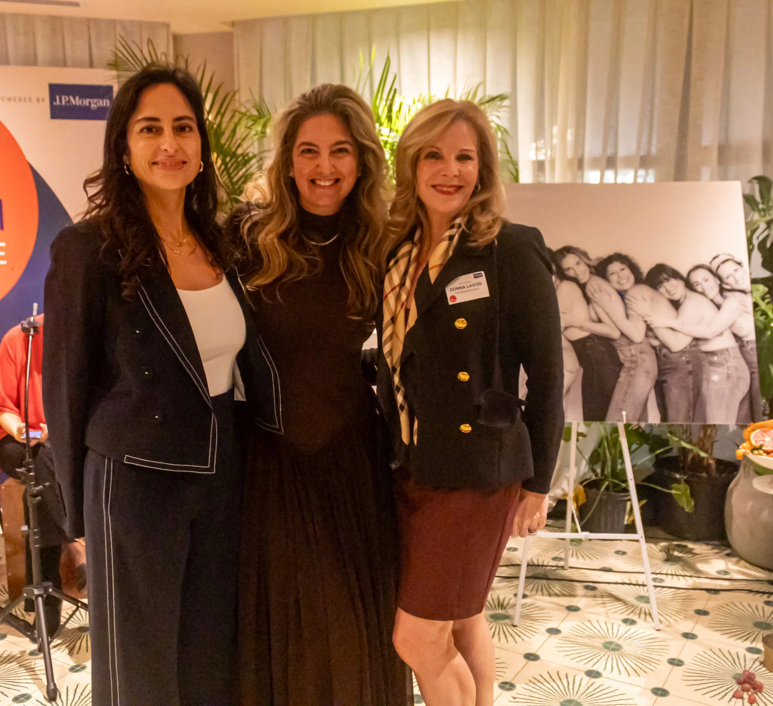 Three women standing together at an indoor event, smiling for the camera. They are standing near a backdrop with a purple and white banner and a collage of photos of women. The woman on the left has dark hair, the woman in the middle has curly gray hair, and the woman on the right has blonde hair. The background includes green plants, a white curtain, and a patterned floor.