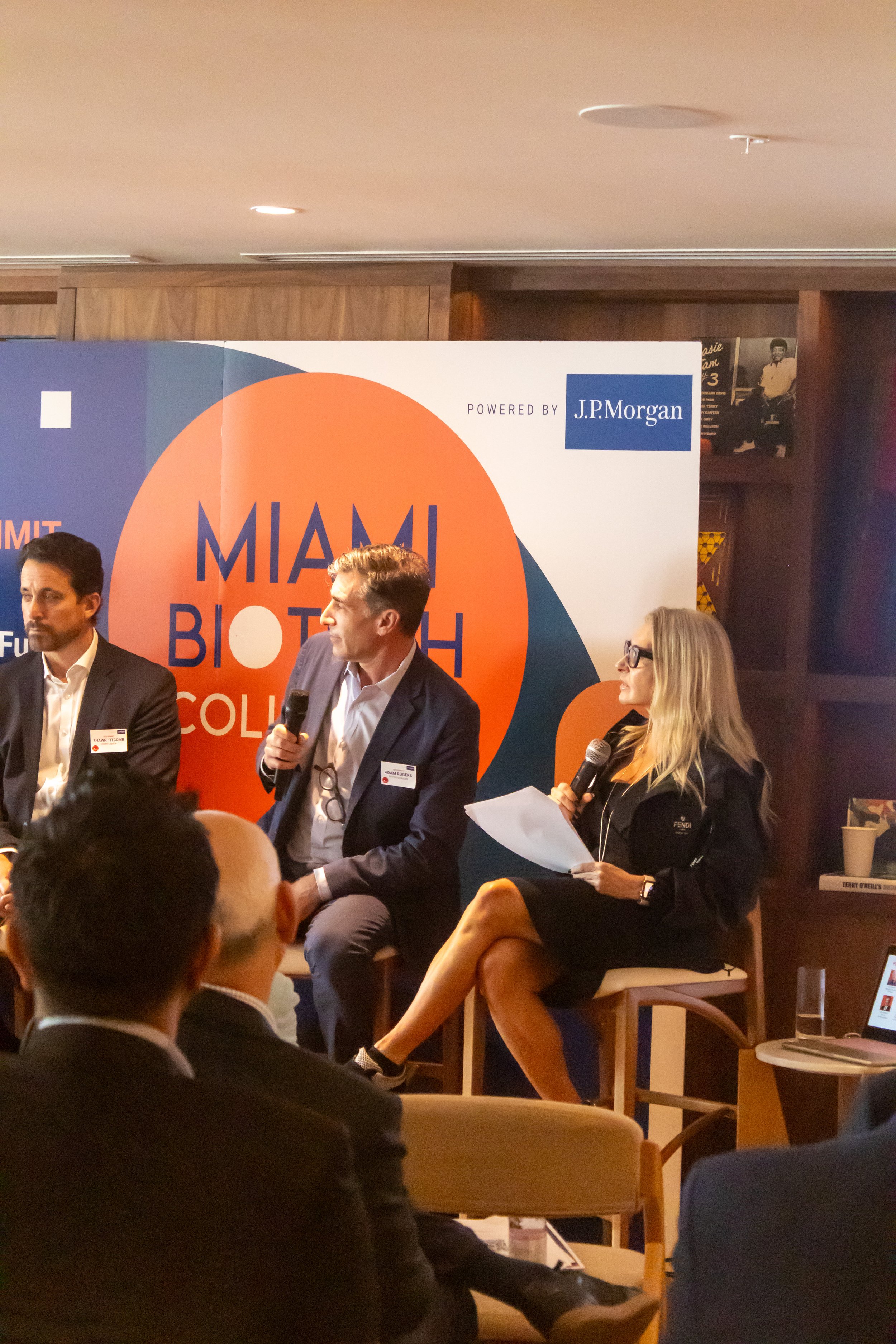 People participating in a discussion panel at the Miami Biotech Collective Summit, with a large sign displaying the event name in the background.