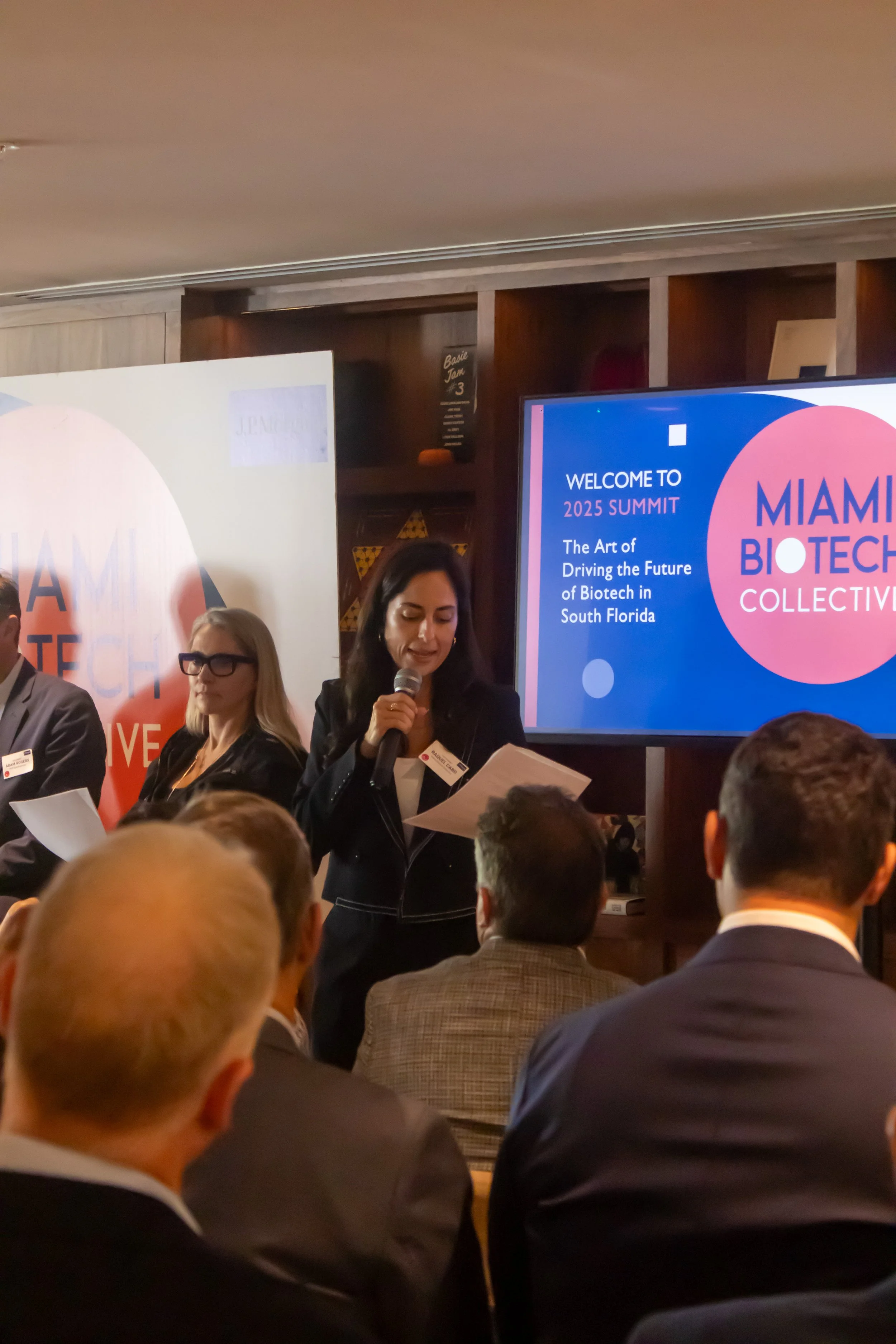 A woman with dark hair speaking into a microphone at a conference, with a large screen displaying the text 'WELCOME TO 2025 SUMMIT,' 'The Art of Driving the Future of Biotech in South Florida,' and the logo for Miami Biotech Collective. Several seated attendees and additional speakers are visible.