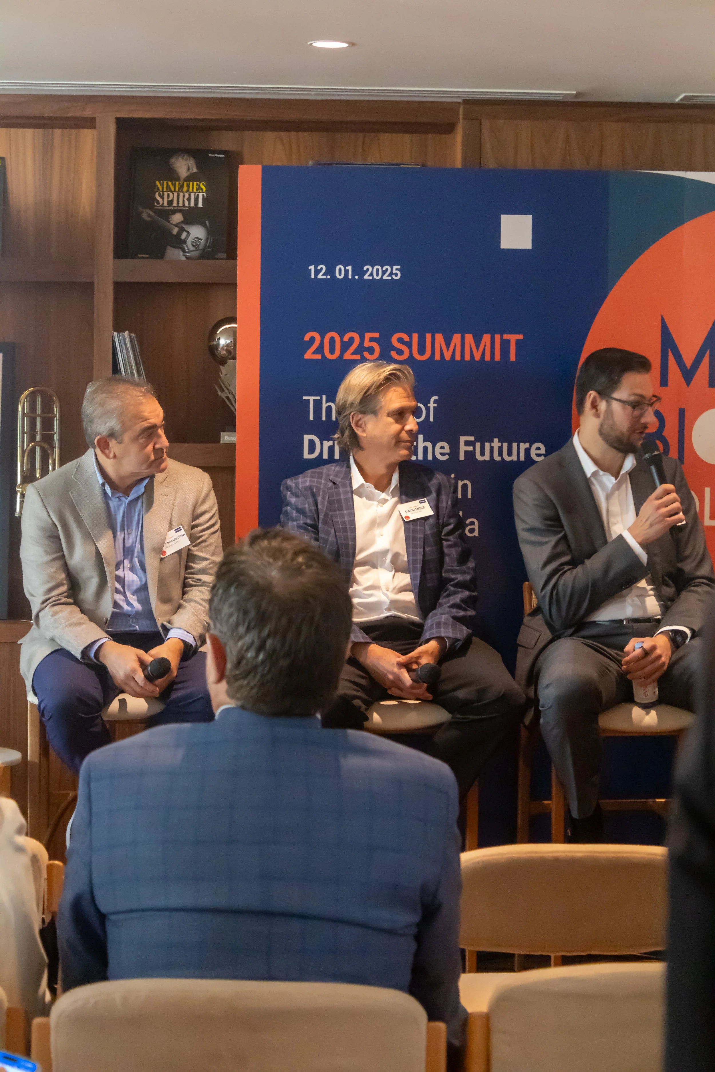 Three men sitting on chairs at a panel discussion or conference, with a blue and orange sign behind them that reads '2025 Summit,' with event details.