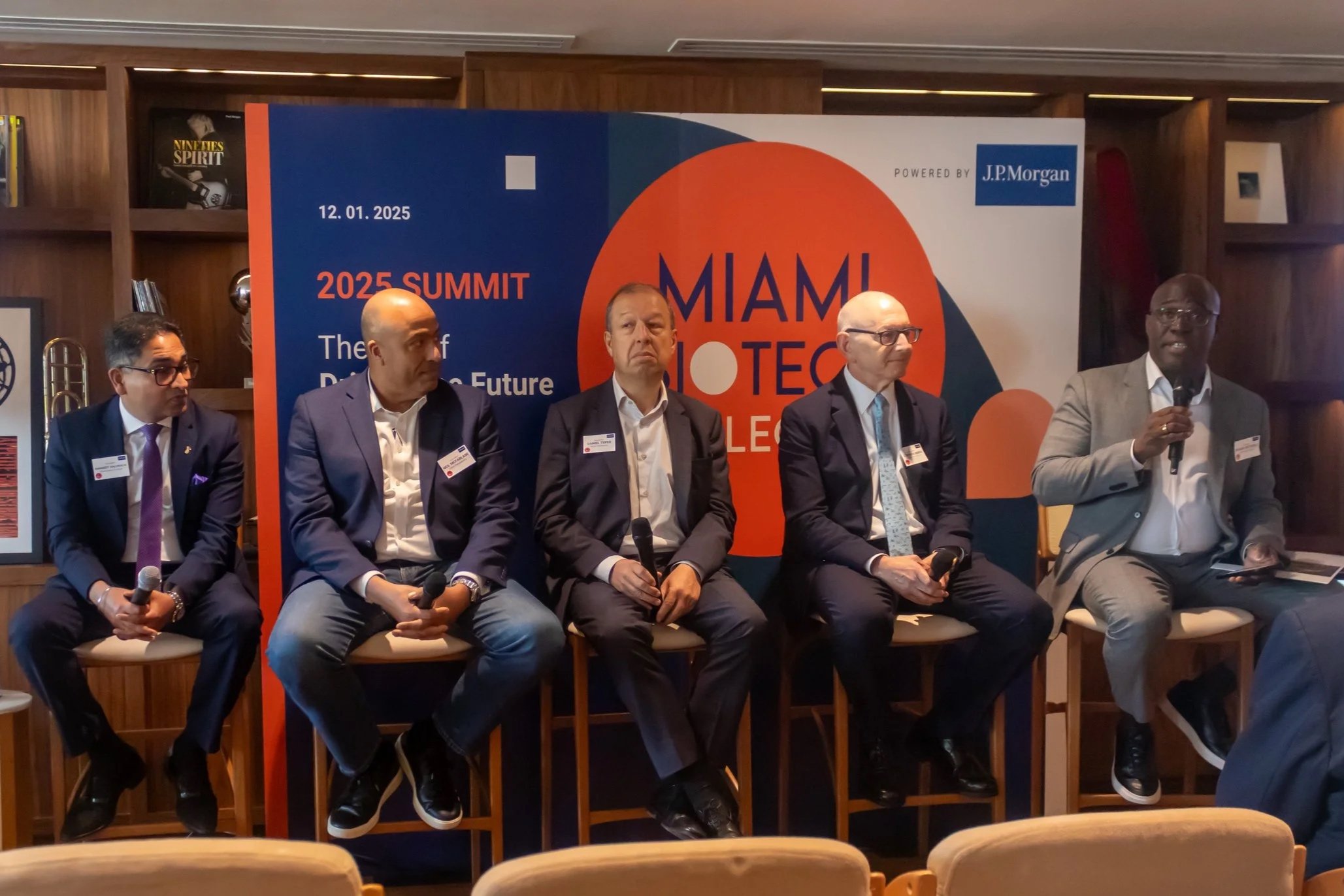 A group of five men sitting on a stage during a panel discussion at the 2025 Miami Biotech Collective Summit, with a large blue, orange, and white banner behind them displaying the event details and the date 12.01.2025.