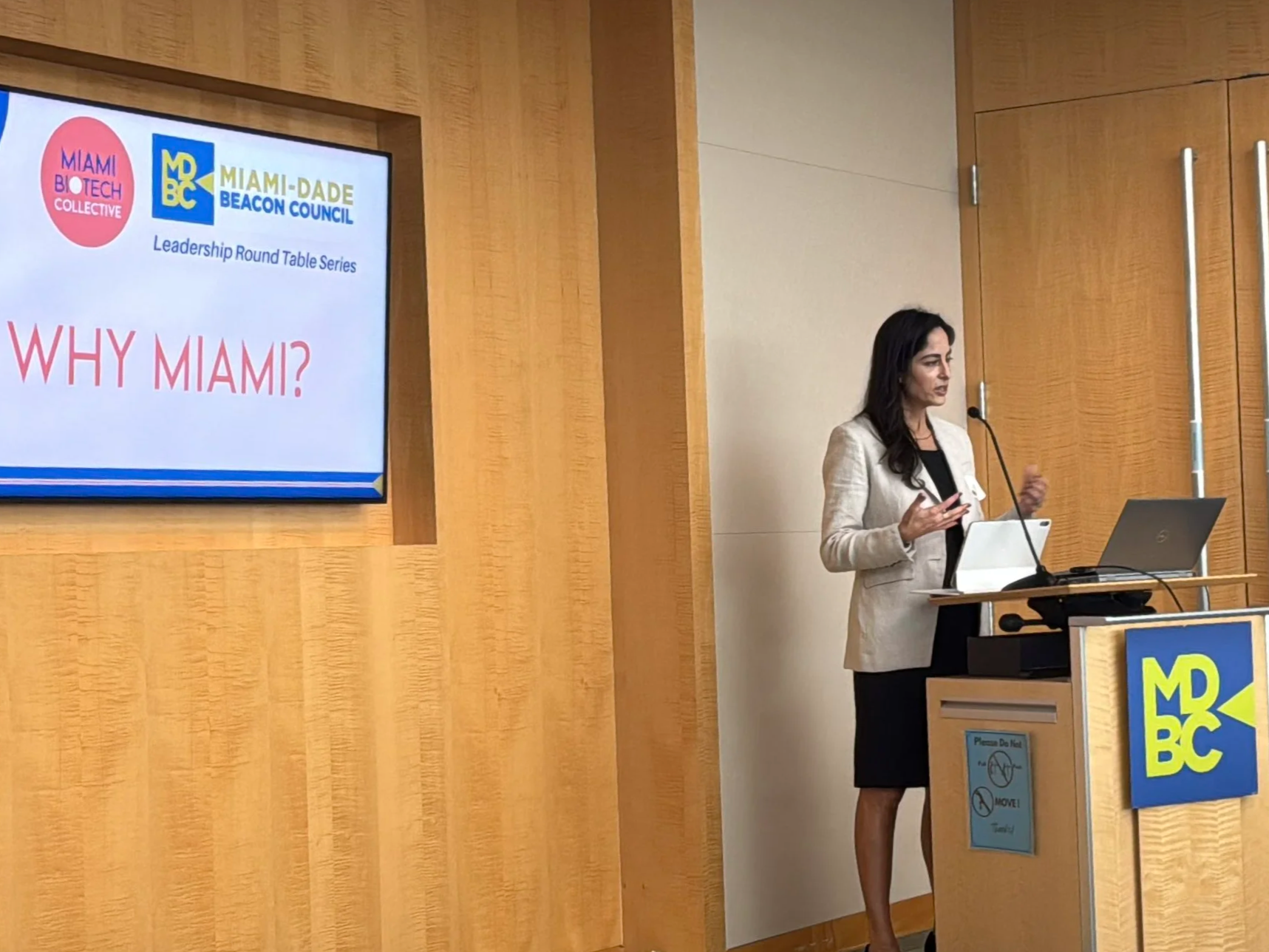 A woman in a beige blazer and black dress giving a presentation at a conference. She stands behind a podium with a microphone, a laptop, and a presentation screen in the background displaying logos and the text 'WHY MIAMI?'