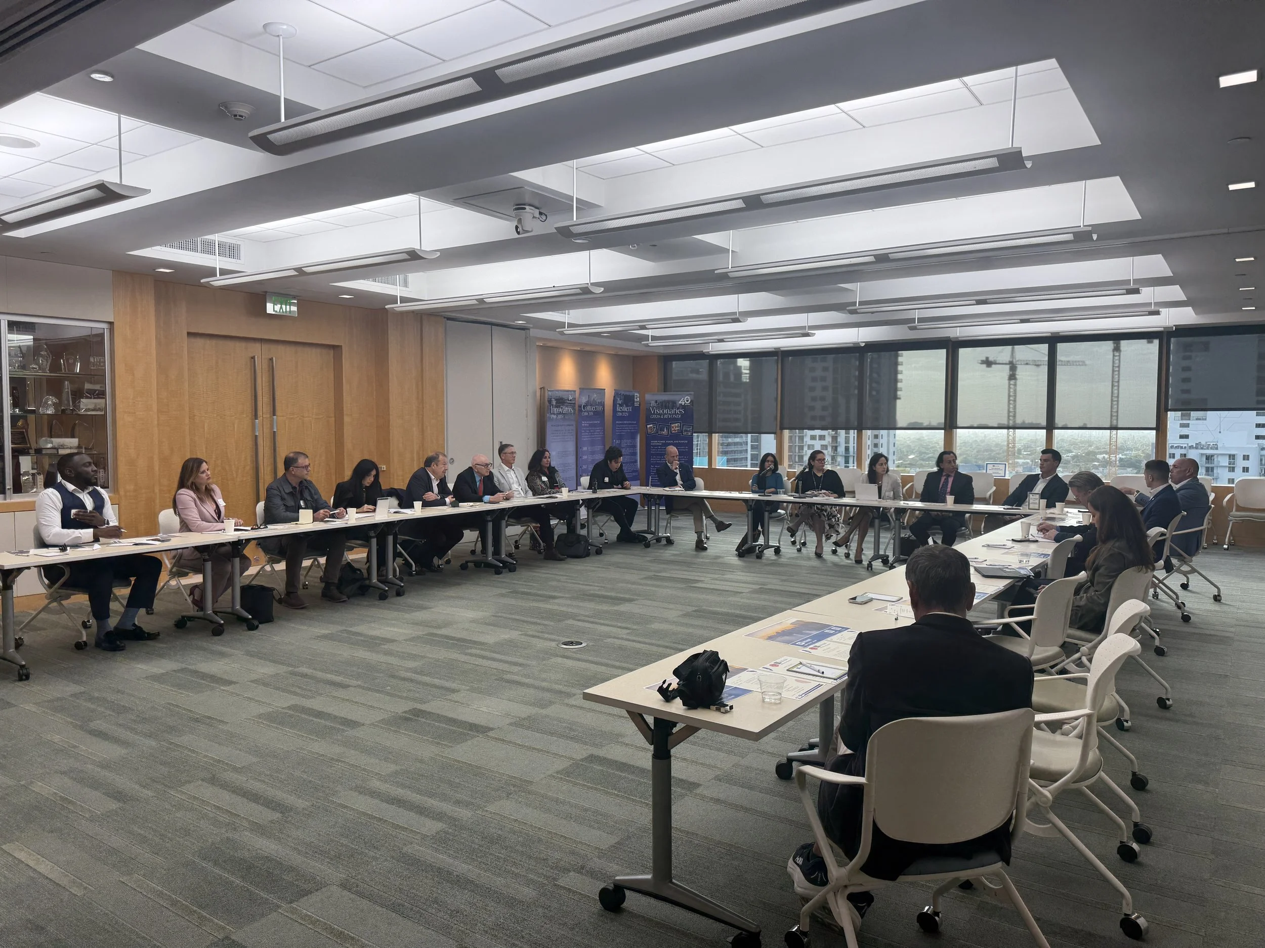 A large conference room with a U-shaped table setup and people seated around it, participating in a meeting. The room has large windows showing a cityscape with construction cranes, and modern ceiling lights.