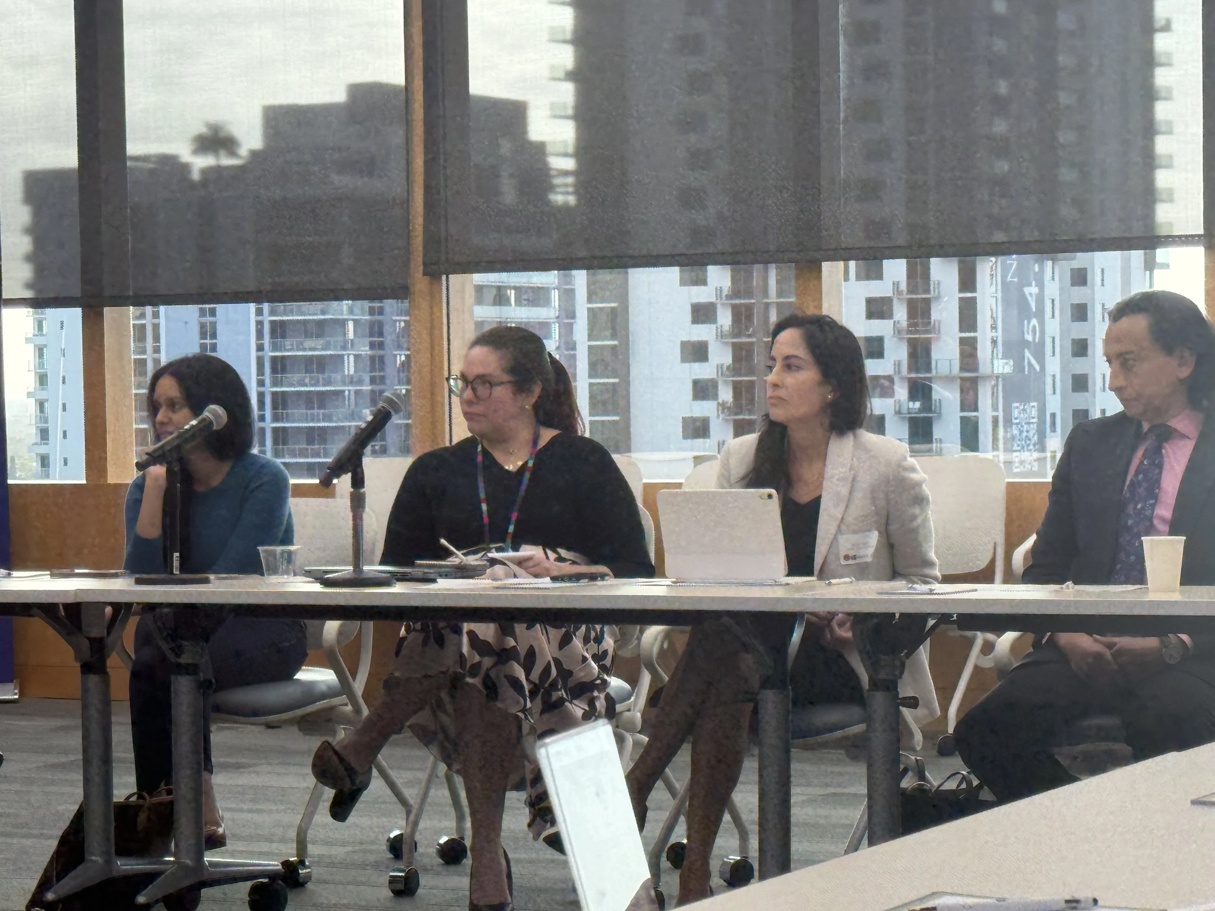 People seated at a table during a panel discussion or conference, with city buildings visible through large windows in the background.