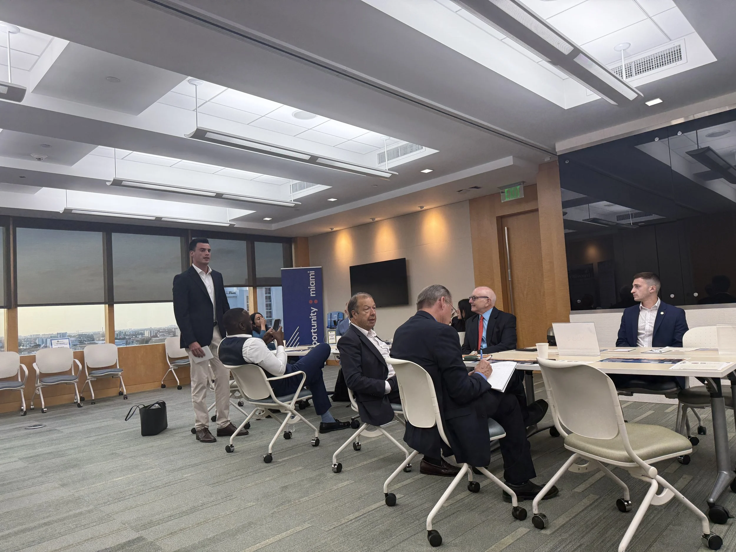 A group of people in business attire sitting and standing around a conference table in a modern office with large windows and ceiling lights.