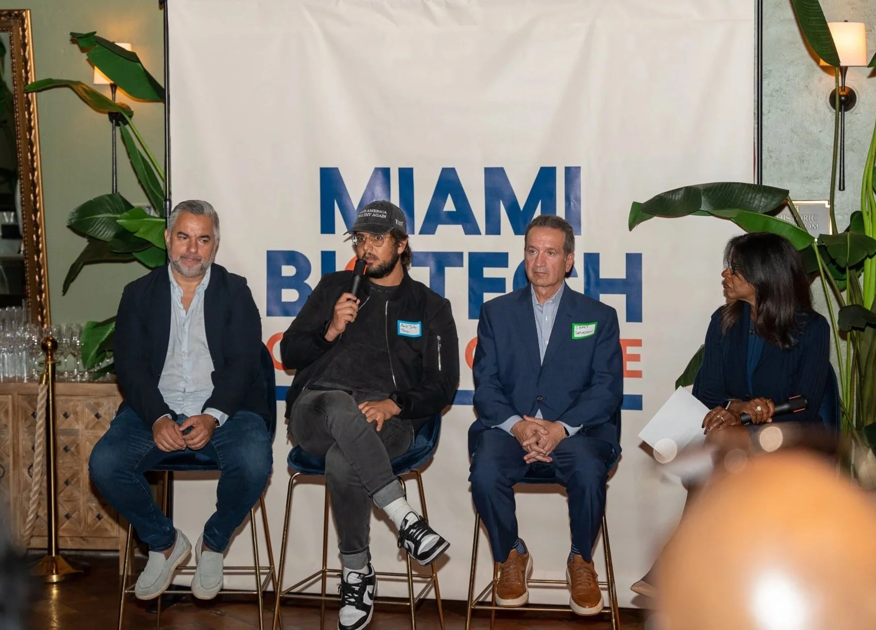 Four individuals seated on stage at a Miami event, with a woman on the far right holding a microphone. Behind them, a large sign reads 'Miami Biotech Collective'. Large green plants and wall lamps are also visible.