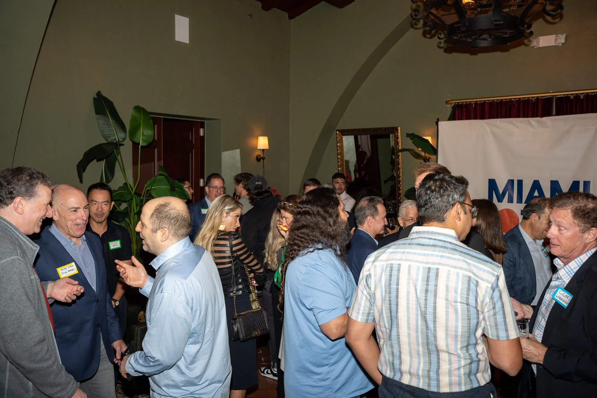 A large group of people gathered indoors at a social or networking event. They are engaging in conversations and wearing name tags. The room has green walls, a large plant, and some wall decorations including a mirror. Attendees are dressed in business casual attire.