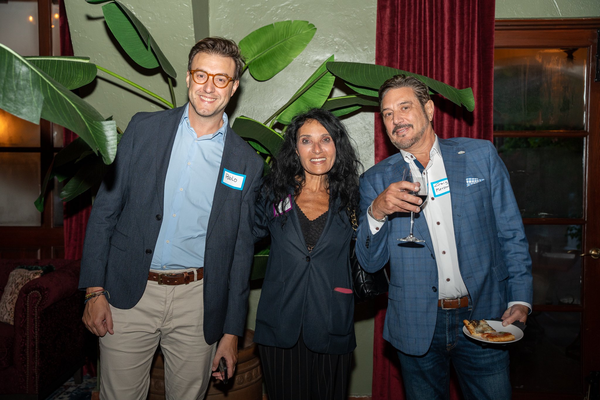 Three people standing together indoors with large green plants behind them. They are dressed in business casual attire, and they are smiling. The man on the right is holding a glass of red wine and a plate with food, and they are wearing name tags.
