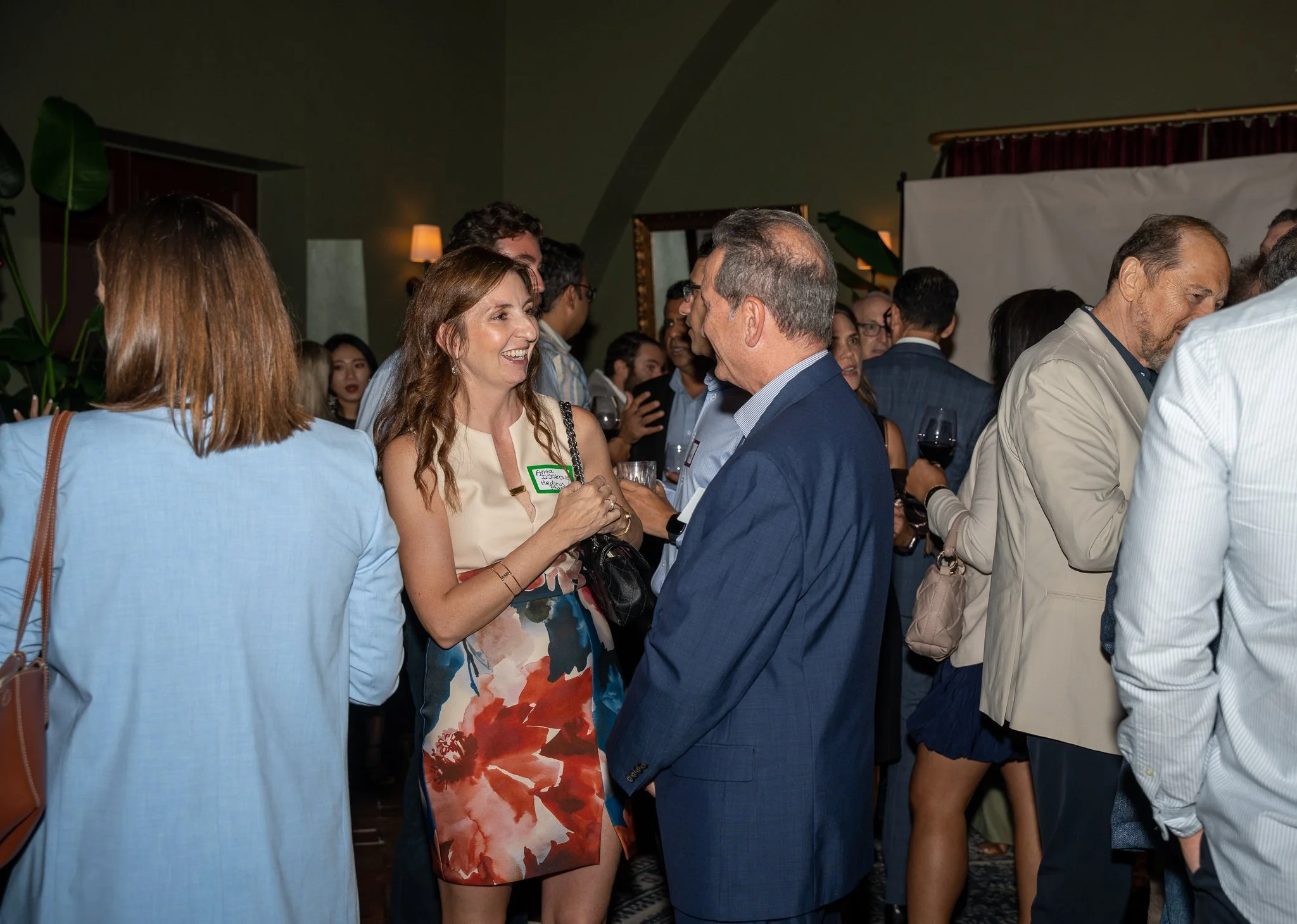 People socializing at a gathering, with two individuals in focus talking and smiling, other attendees in the background, in an indoor setting with dim lighting and decorative elements.