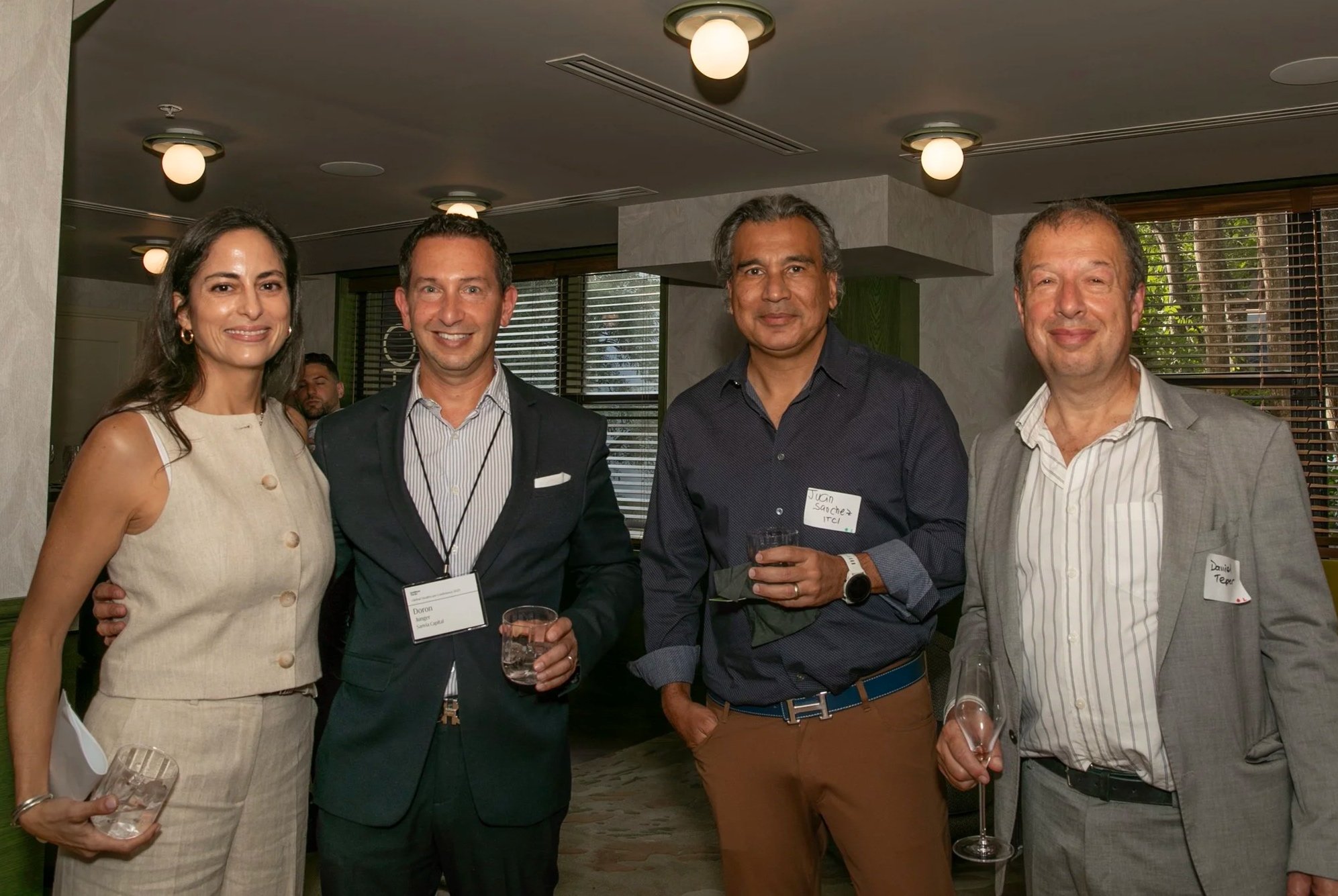 Four people are standing indoors at a social event, smiling at the camera. They are dressed in business casual to formal attire, each holding a glass of drink. The background shows a lit ceiling with round lights and windows with blinds.