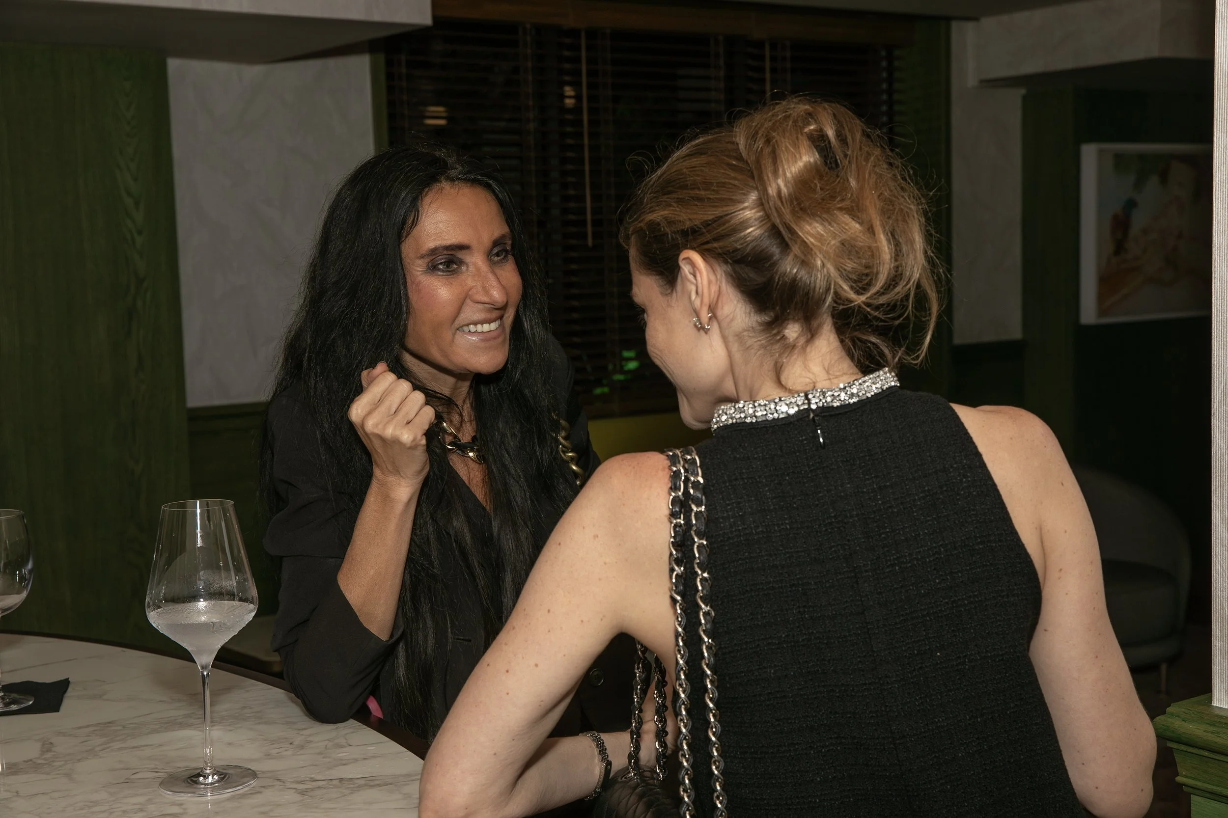 Two women engaged in conversation at a bar or restaurant, with wine glasses on the table in front of them.