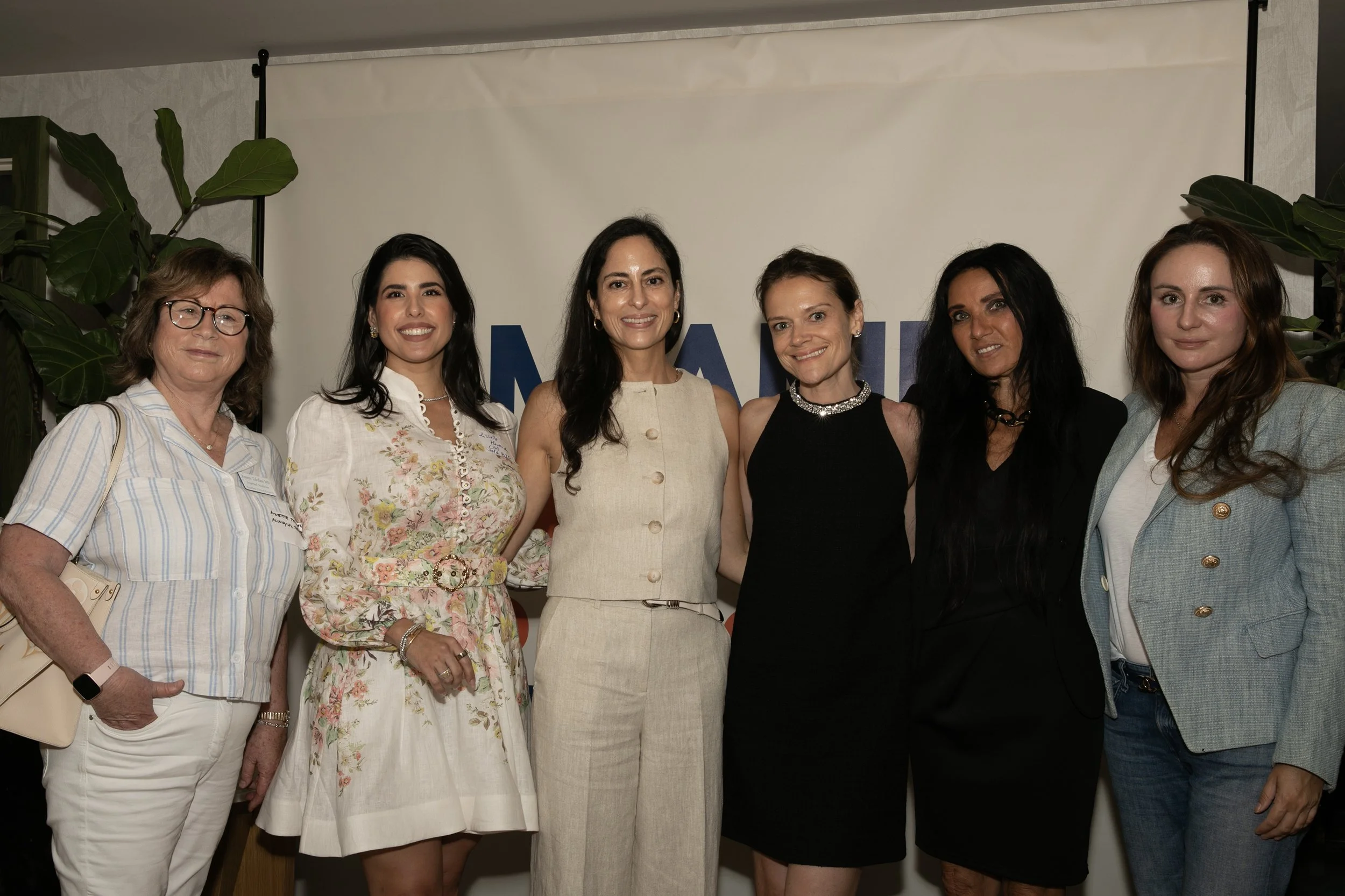 A group of seven women standing together indoors in front of a white background with large green plants on each side.