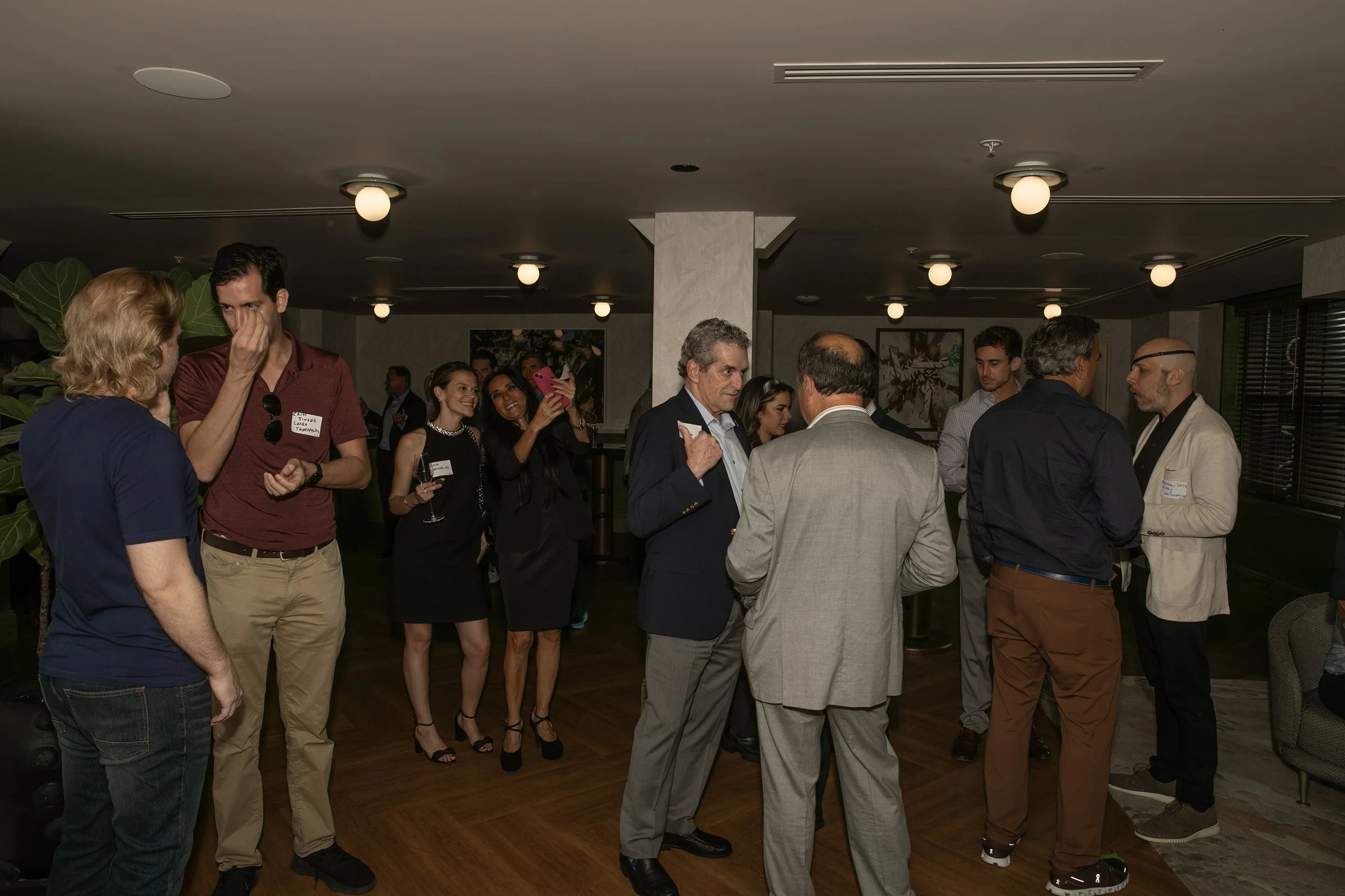 Group of people socializing at a party or networking event indoors, some holding drinks, engaging in conversation and taking photos.
