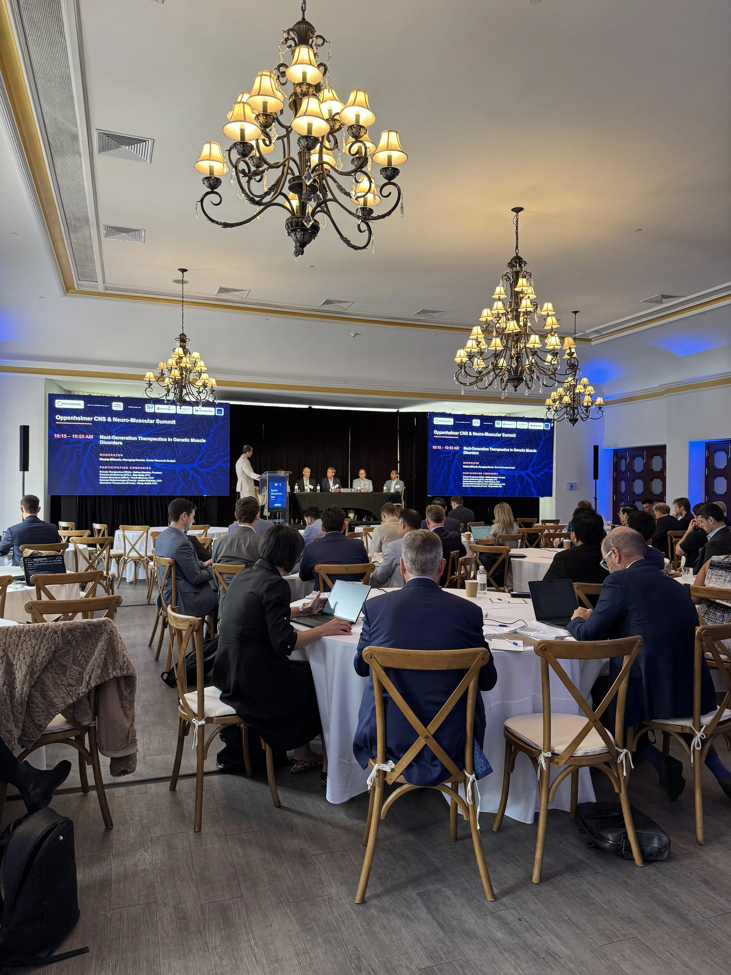 A conference room filled with attendees seated at round tables, listening to a panel of speakers on stage. Large screen displays conference information, and ornate chandeliers hang from the ceiling.