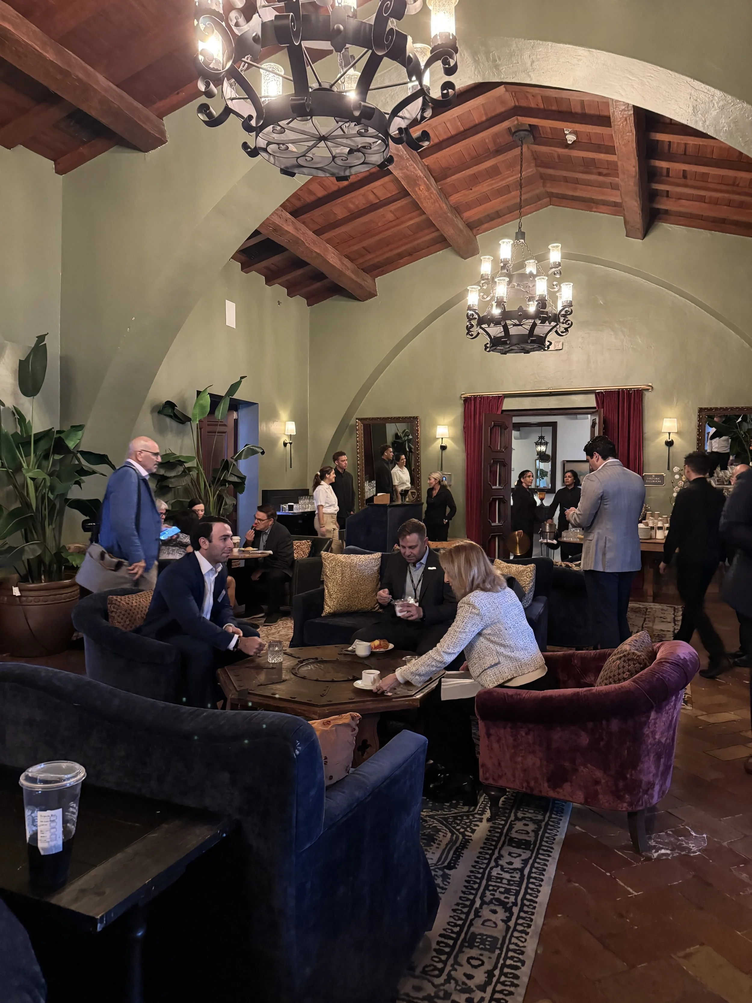 An elegant restaurant interior with green walls, wooden beams, chandeliers, and multiple people dining and socializing.