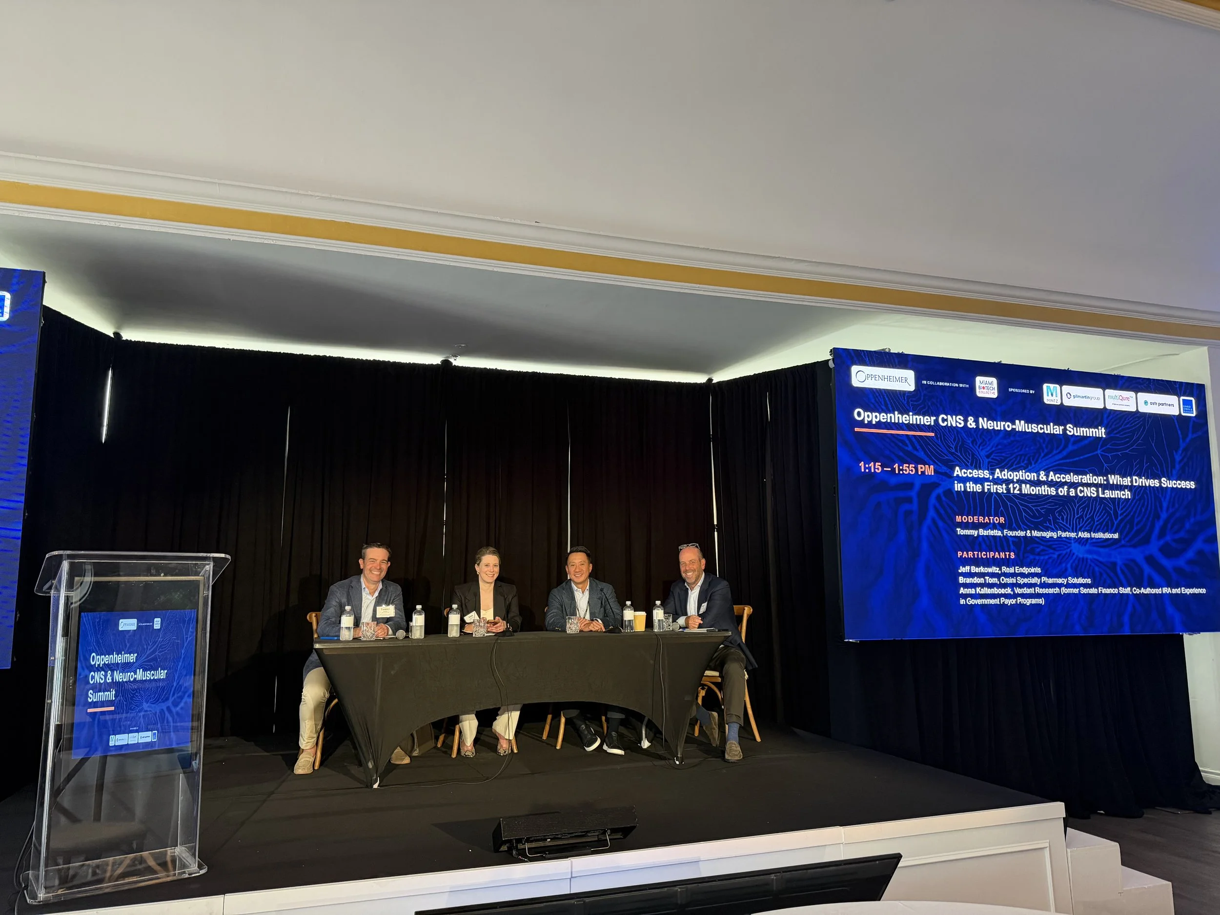 Four people seated at a panel table on a stage at a medical conference, with a blue conference banner behind them displaying the title "Oppenheimer CNS & Neuro-Muscular Summit" and scheduled session info.