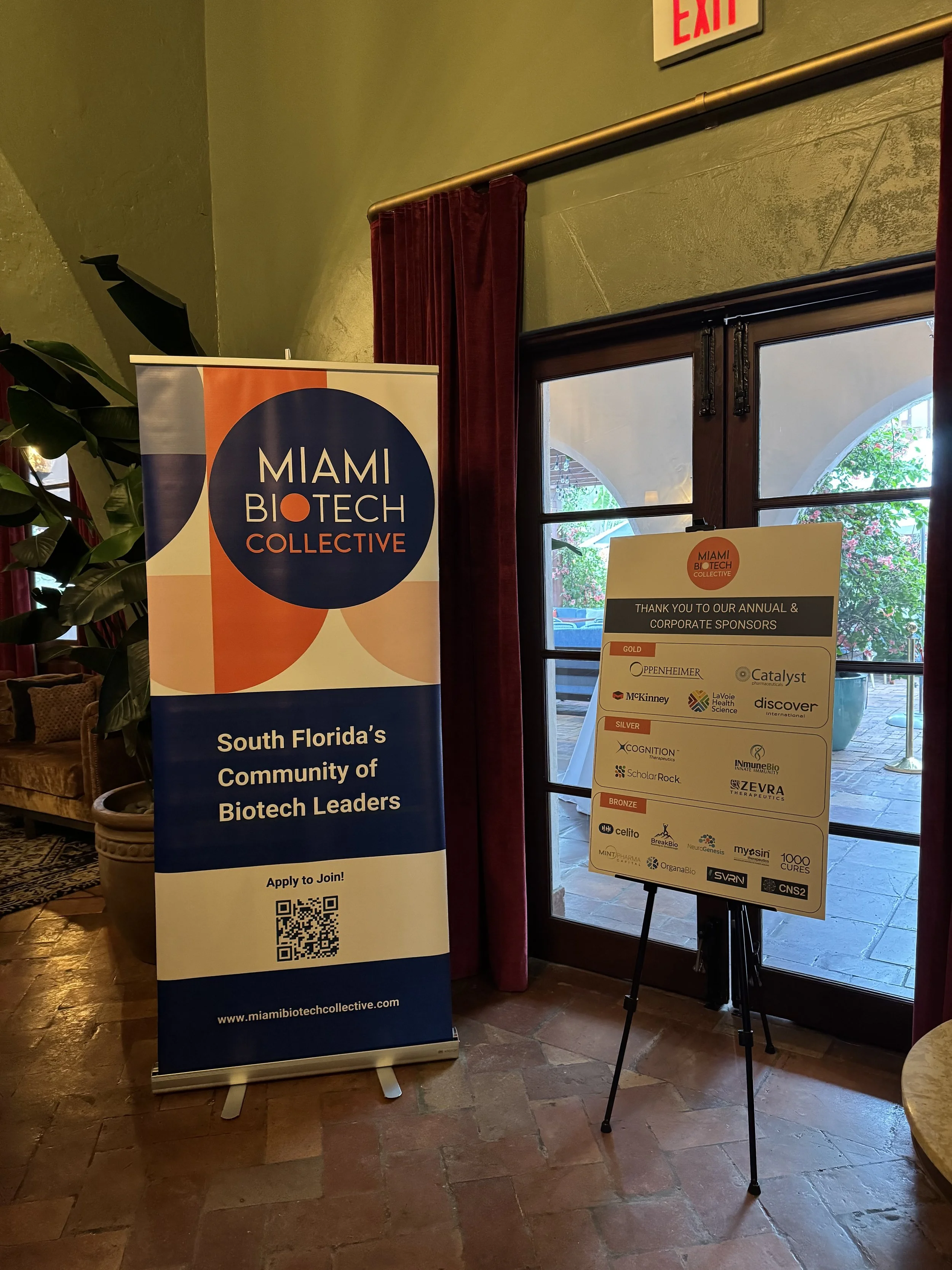 Indoor event with two signs promoting Miami Biotech Collective; one vertical banner with logo, website, and QR code, and a smaller informational board listing sponsors, all near a window and wooden door