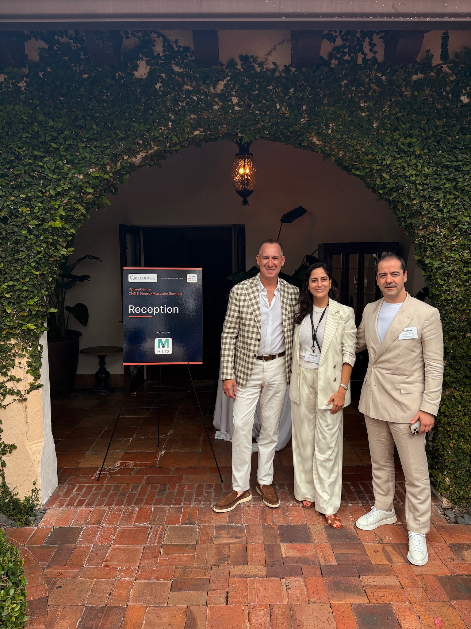 Three people standing under a green leafy archway at an event, with a reception sign and decorative lighting in the background.