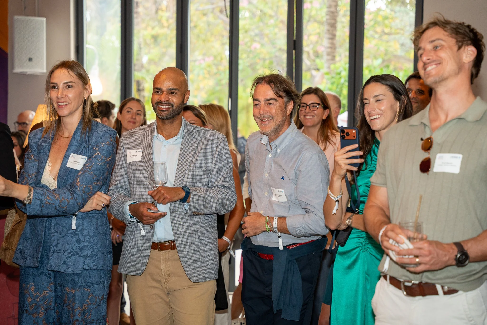 A group of diverse people attending a social event or conference indoors, smiling and talking, with large windows showing trees outside.