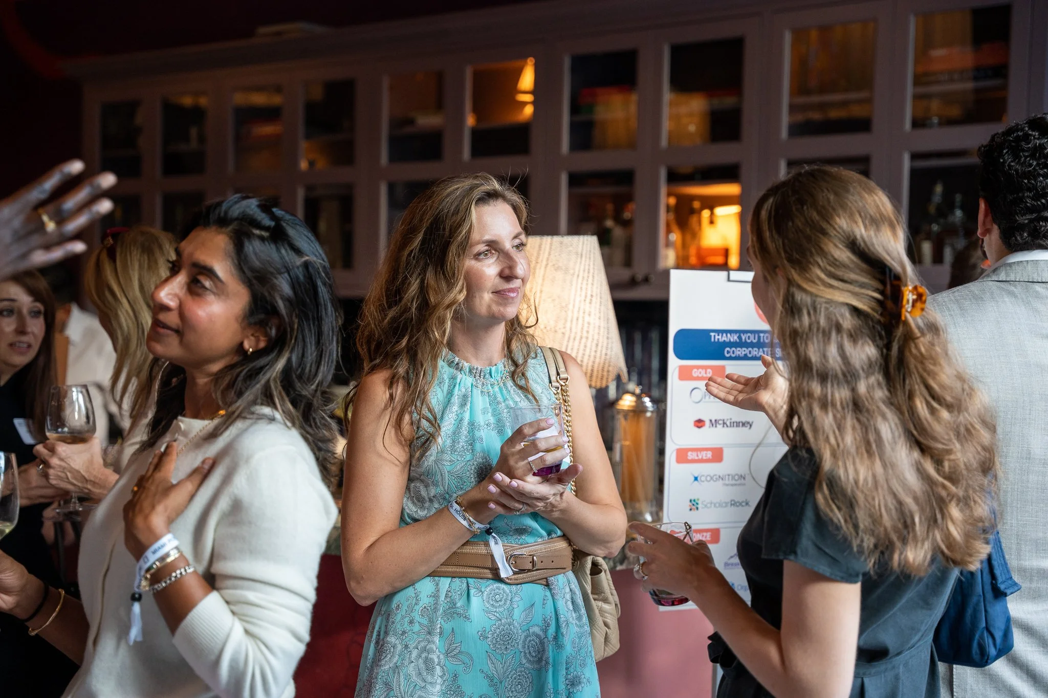 Group of people at a social event, engaging in conversations, with a woman in a blue dress holding a glass of wine in the center. Background features a wooden bookshelf and a display board.