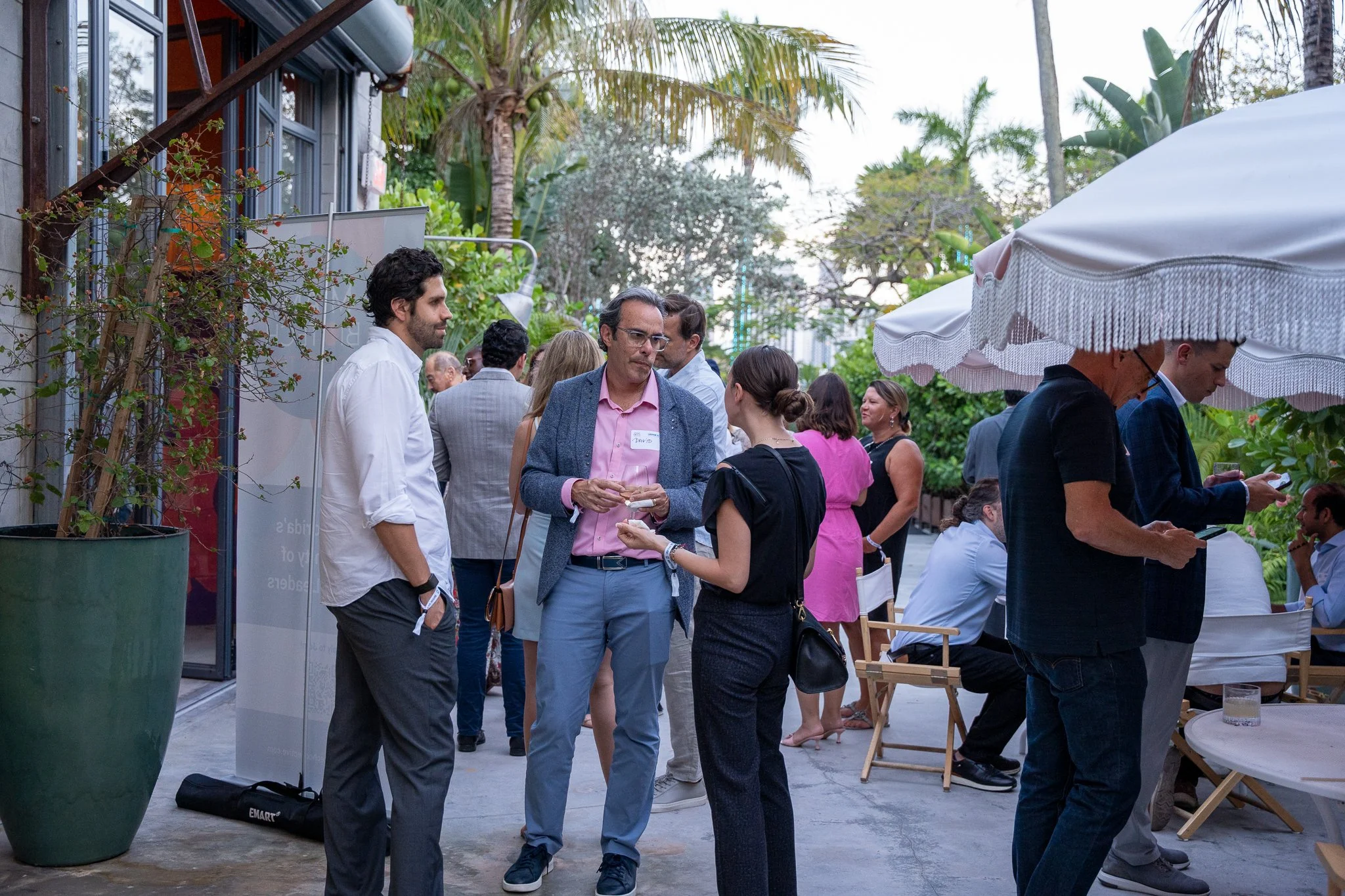 People gathered outdoors at a social event, engaged in conversation, with a mix of men and women dressed in business casual attire, surrounded by greenery, large potted plants, and a white umbrella.