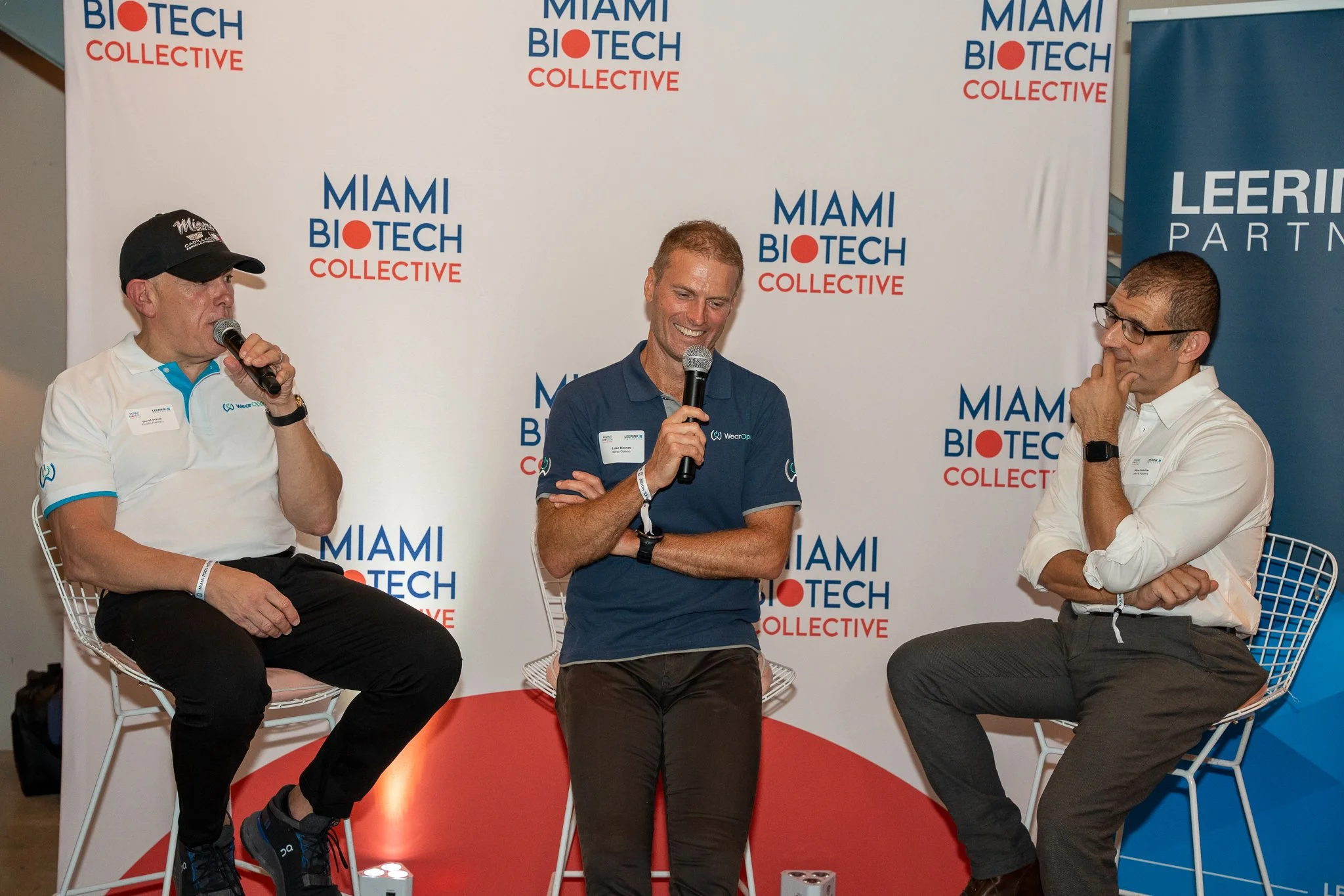 Three men sitting on chairs on a panel at Miami Biotech Collective event. The background displays Miami Biotech Collective logos. One man is speaking into a microphone, another man is smiling, and the third man is listening with his hand on his chin.