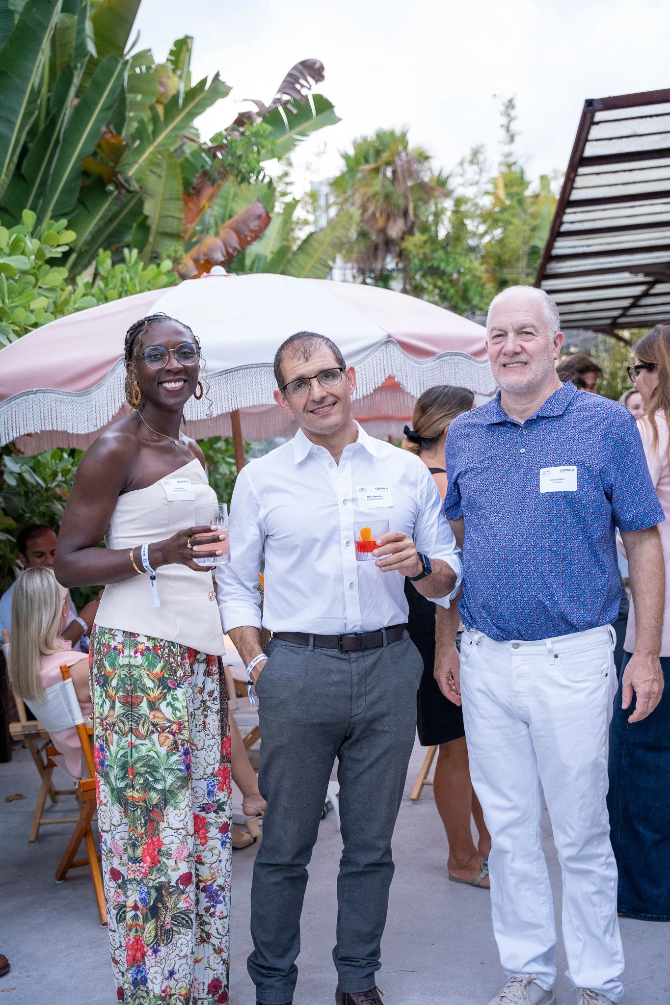Group of three people standing outdoors at a social event, with tropical plants and other attendees in the background. The woman on the left is wearing glasses, gold jewelry, and a strapless white top with a floral skirt, holding a glass of wine. The man in the middle is wearing glasses, a white shirt, and gray pants, holding a cocktail. The man on the right has a beard, is bald, and is wearing a blue patterned shirt with white pants.