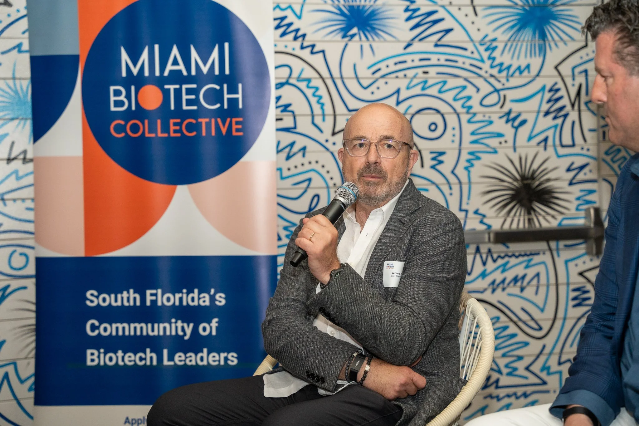 A man with glasses and a gray blazer speaking into a microphone at a conference, with a blue and white patterned background and a Miami Biotech Collective banner.