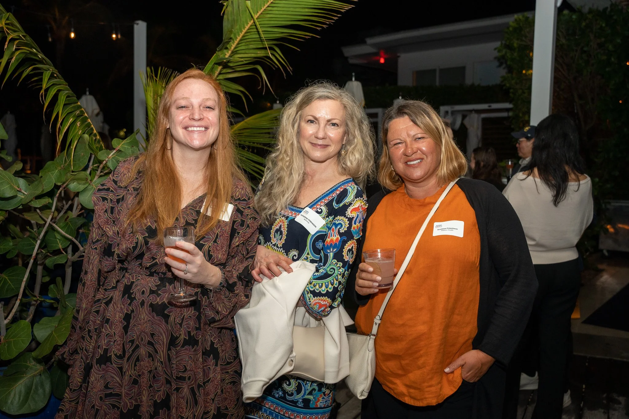 Three women smiling and holding drinks at an outdoor evening gathering, with palm leaves and other guests in the background.