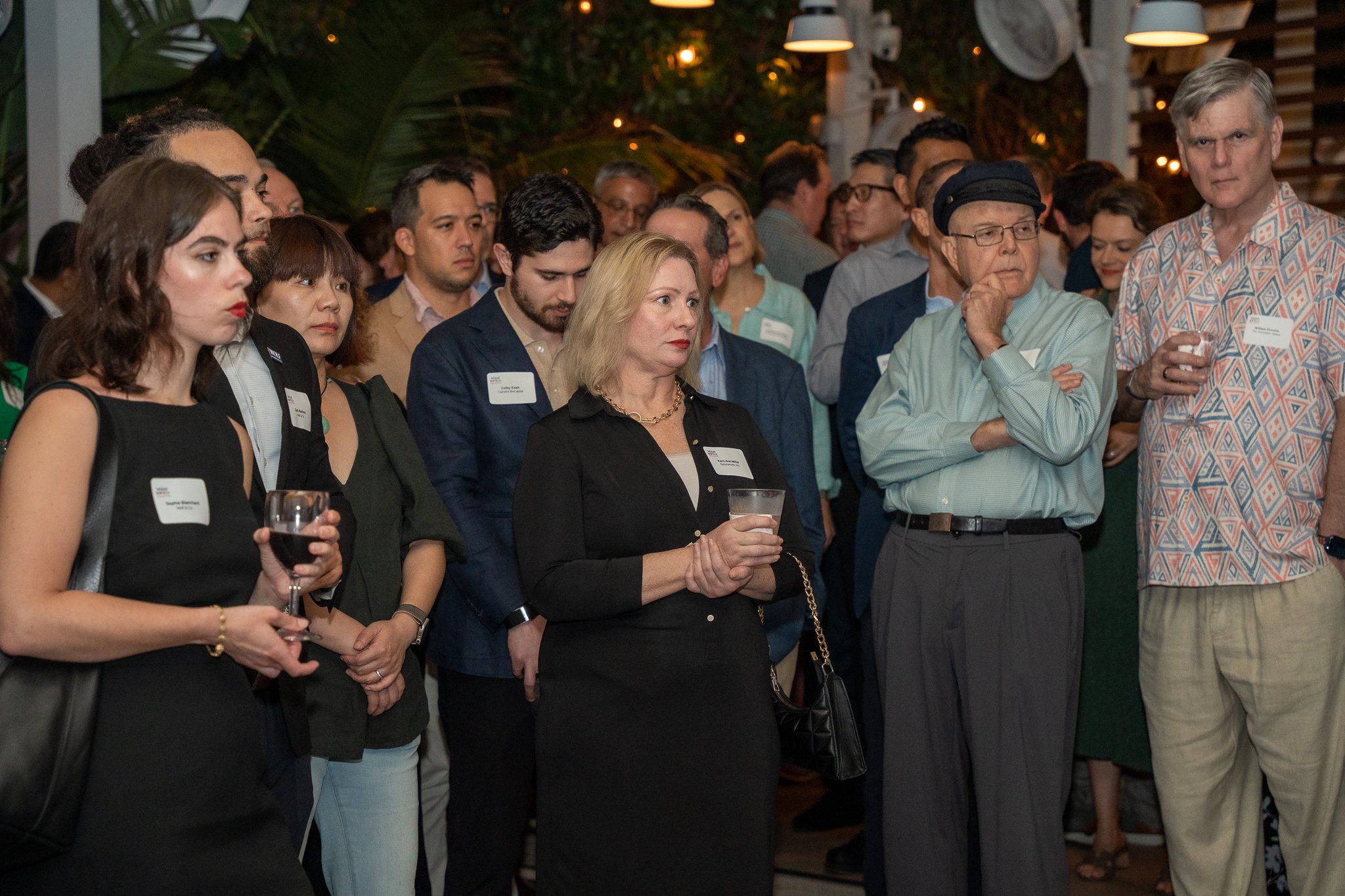 A group of diverse people attending an indoor social event. Some are holding drinks and wearing name tags. The setting includes hanging lights and a leafy, decorative background.