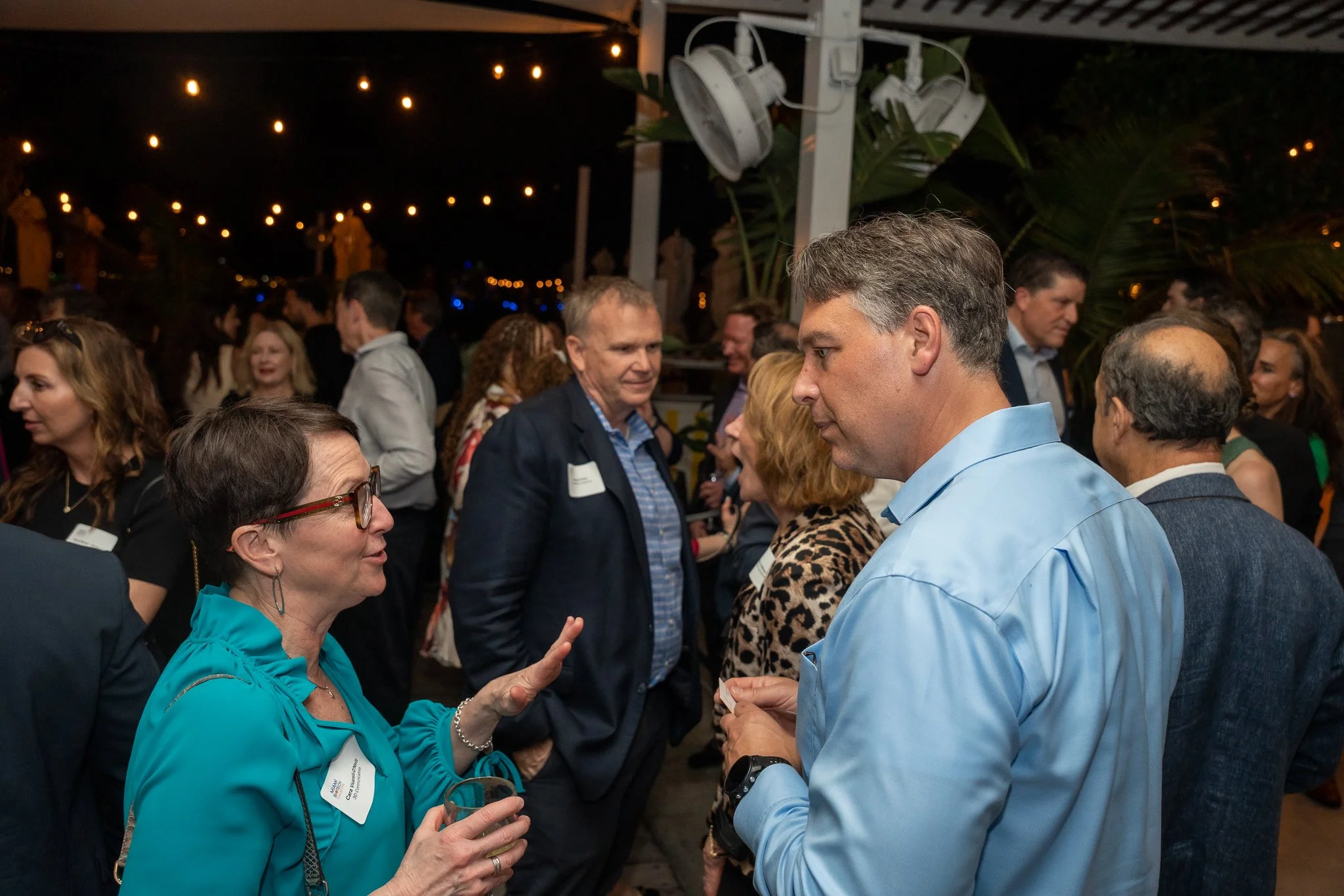 A woman in a teal blazer and glasses is talking to a man in a light blue shirt, holding a glass of wine, at a social gathering, with multiple people engaging in conversations in the background under string lights.