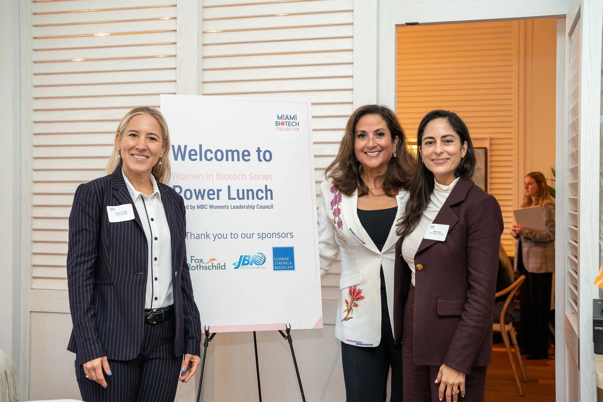 Three women standing together at an indoor event, smiling. Behind them is a welcome sign for a women's biotech series power lunch, with sponsor logos at the bottom.