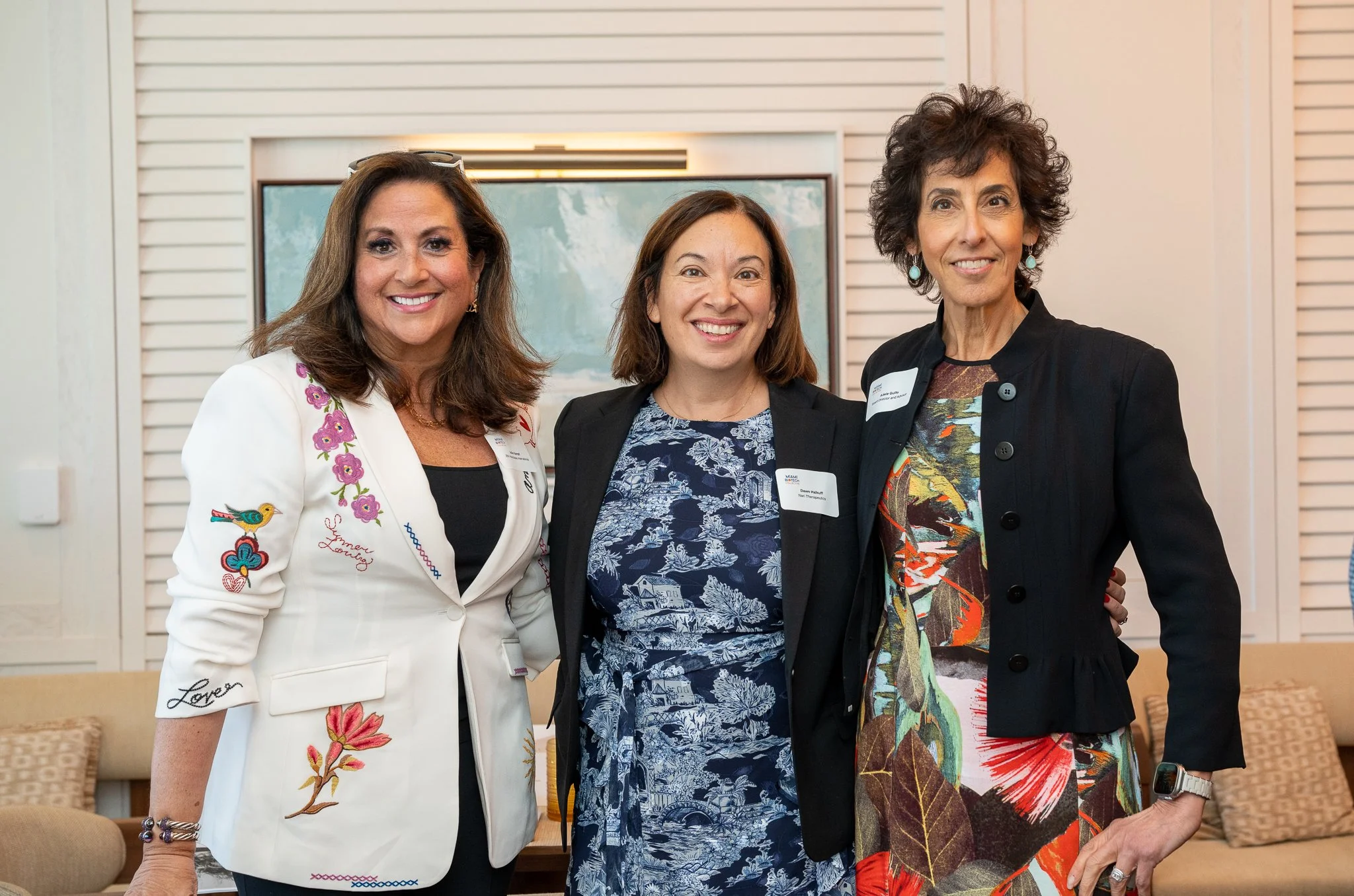 Three women standing close together, smiling, in an indoor setting with a painting on the wall behind them.