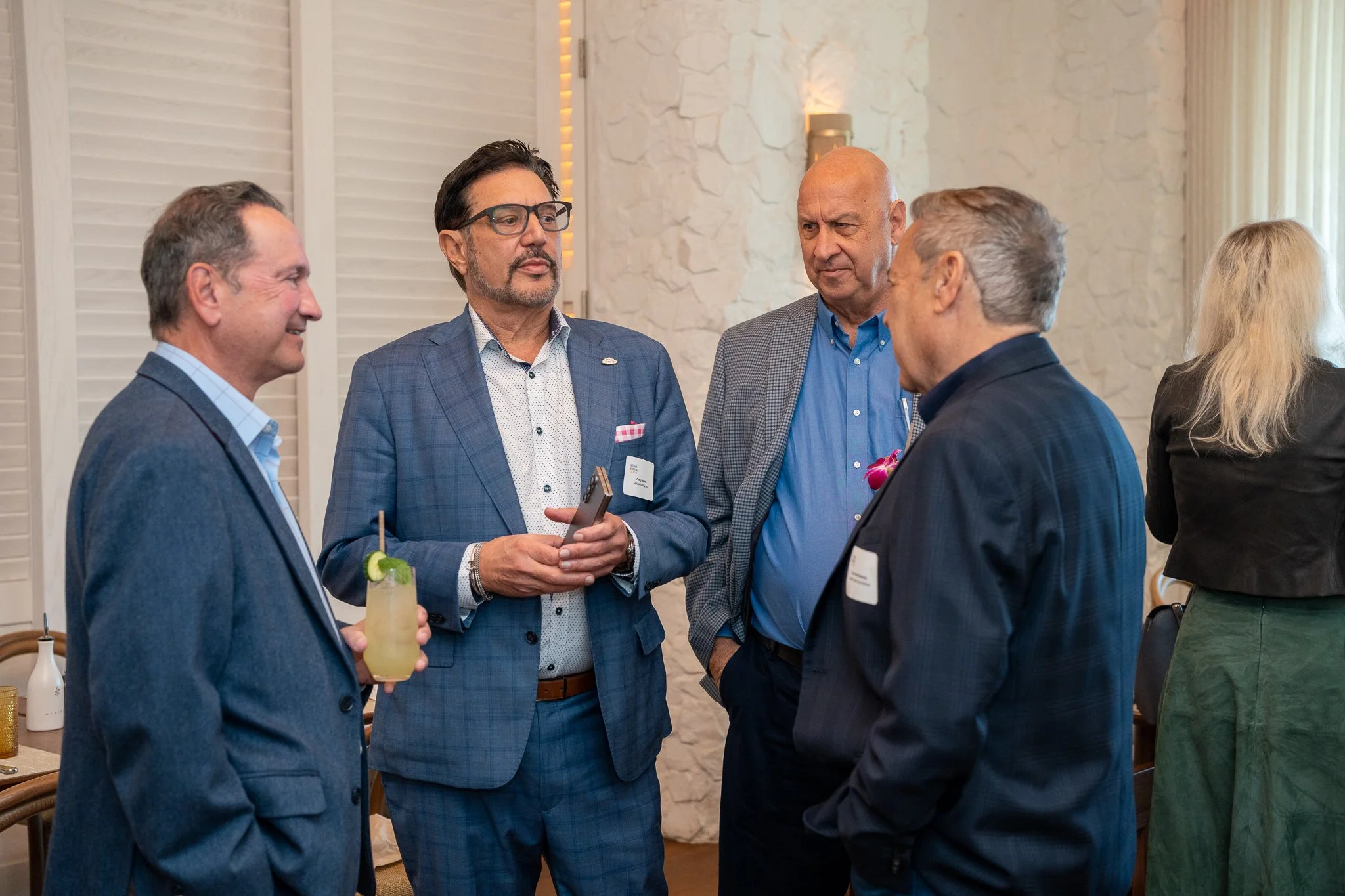Group of five adults in conversation at a social event, one holding a drink, in a well-lit room with white brick walls.