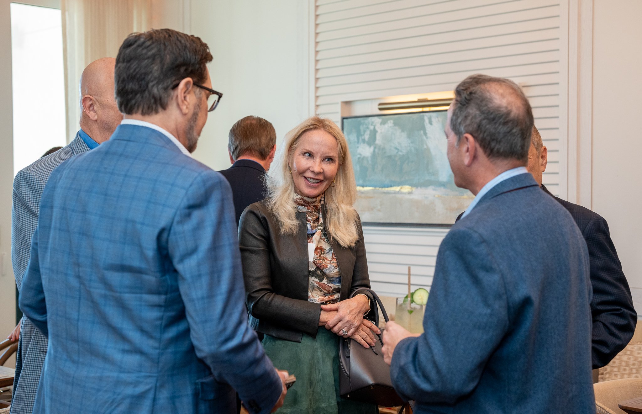 A group of professionals engaged in conversation at a business or networking event, wearing formal attire and standing in a well-lit room with a large painting in the background.