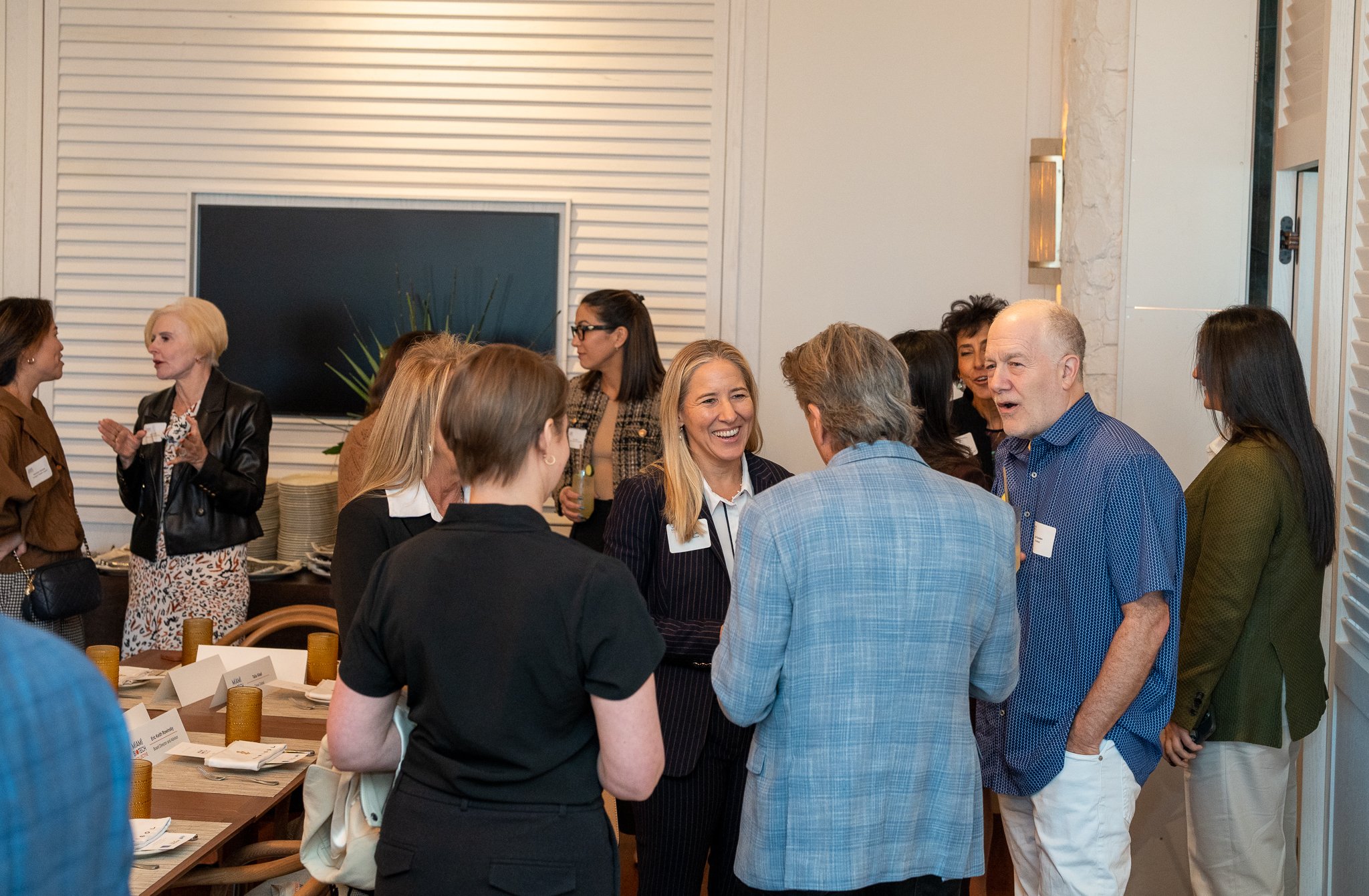 Group of people engaged in conversation at a social event in a modern indoor setting, with a white wall, chalkboard, decorative plants, and tables with place cards and glasses.